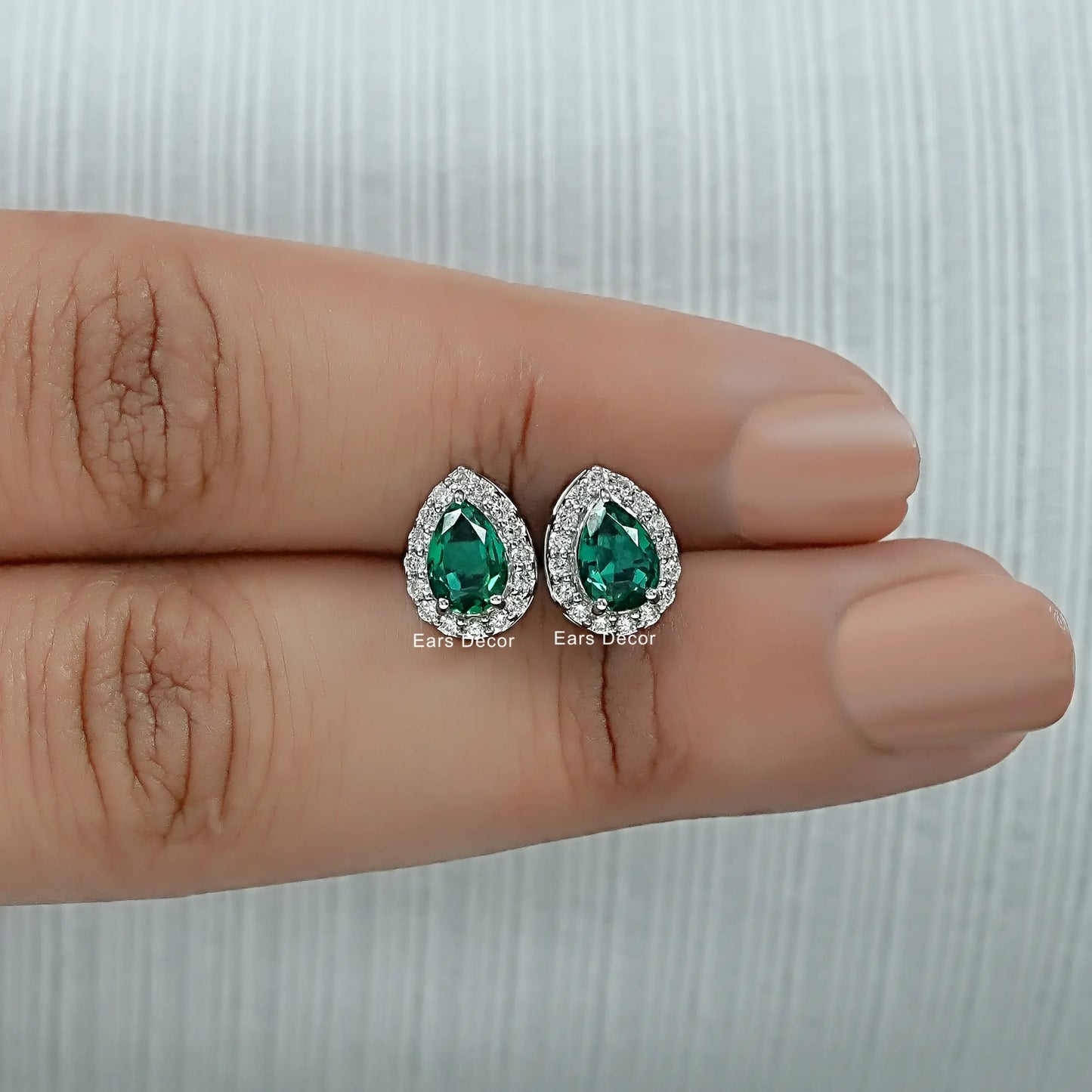 Dainty pear shaped emerald stud earrings with lab diamond halo held between two fingers size reference nude nails close-up