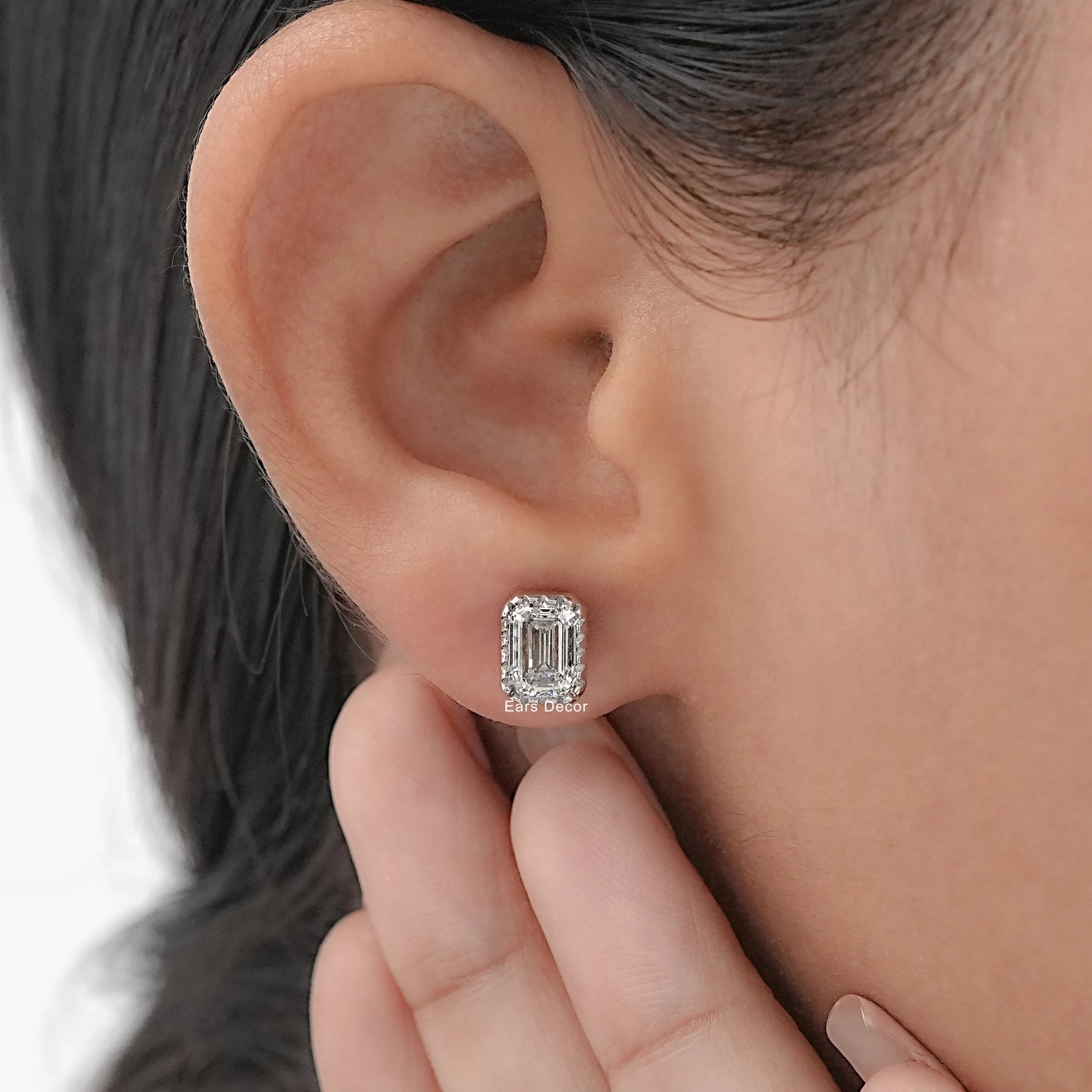 Emerald cut diamond stud earrings in bezel setting worn on model's ear fingers visible