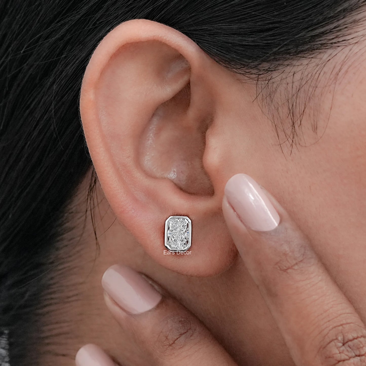 Octagon shaped bezel moissanite solitaire earring worn on model ear, nude nails fine jewelry
