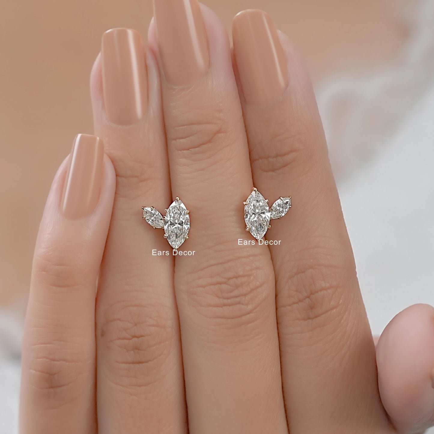 Marquise and pear cut diamond earrings with prong setting held between the fingers of a hand with nude nails.