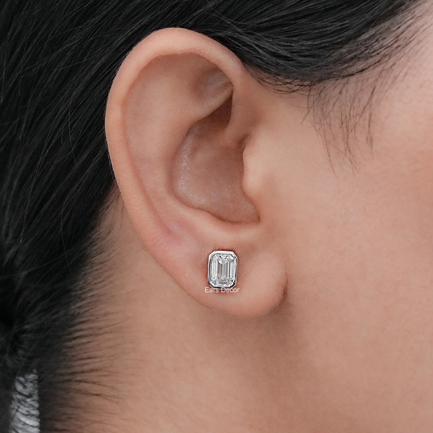 Dainty emerald cut moissanite diamond stud earrings white gold bezel worn model ear dark hair warm skin side profile close-up