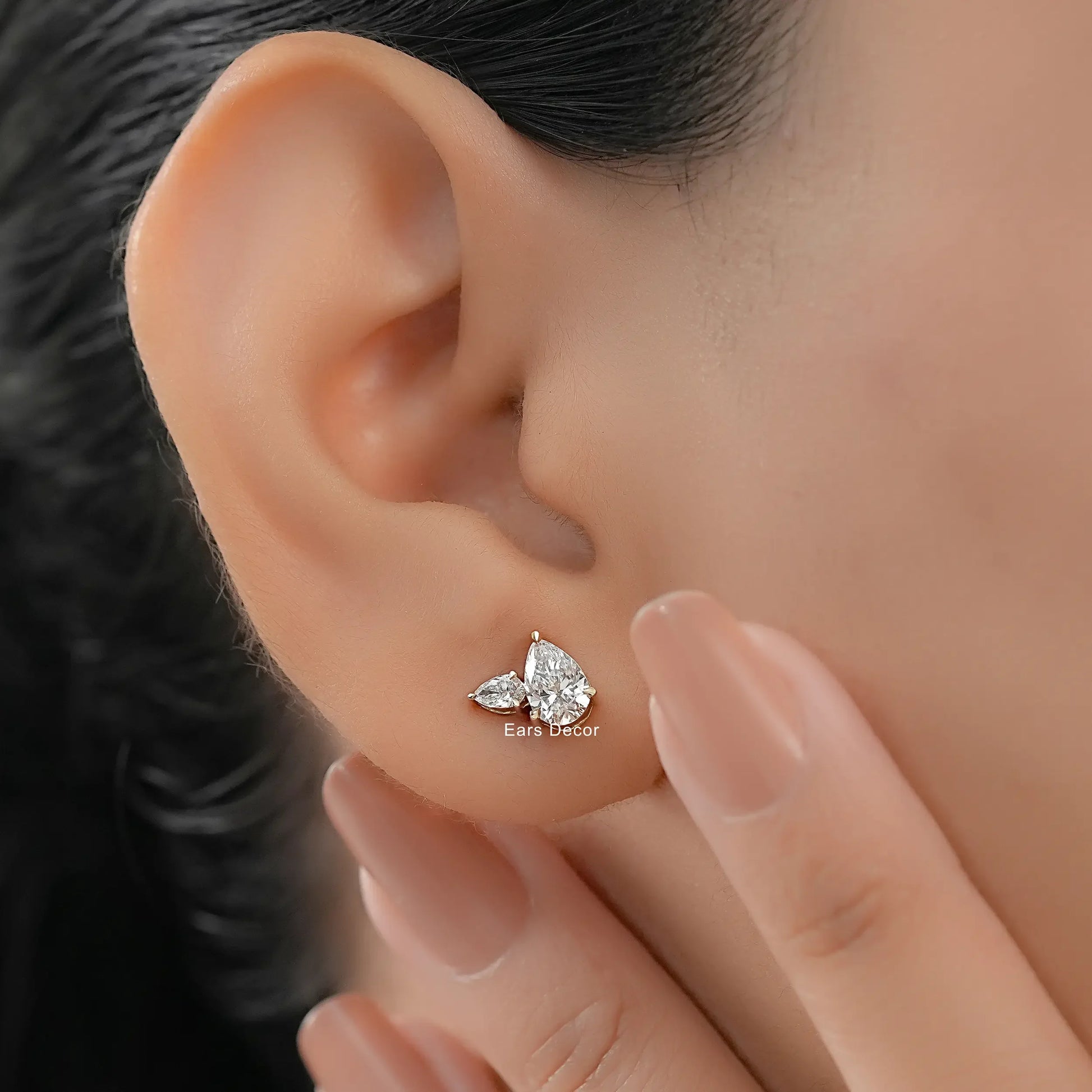 Small pear cut diamond stud earrings solid yellow gold worn on model ear close-up dark wavy hair nude pink nails hand touching cheek