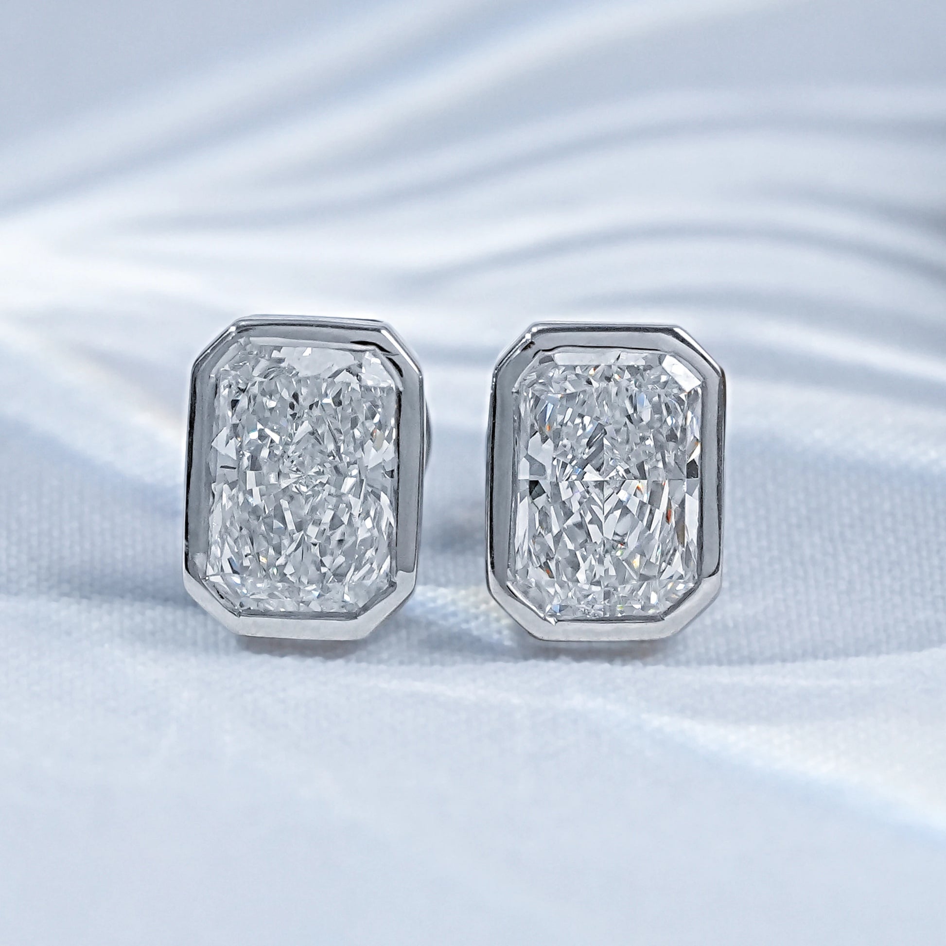 Radiant cut diamond bezel stud earrings pair white gold on satin flatlay close-up