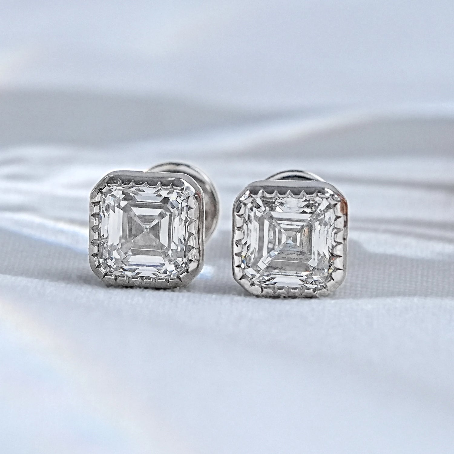 A magnificent pair of asscher milgrain lab diamond stud earrings held between the fingertips, showcasing the intricate beaded border and vintage inspired step cuts.