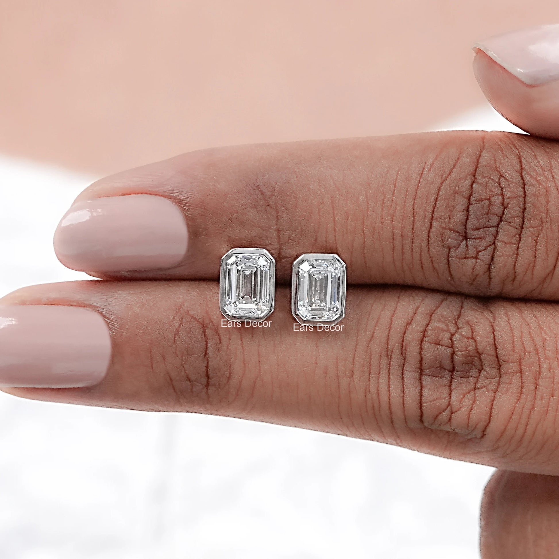 Classic emerald cut diamond stud earrings white gold bezel pair displayed on palm dark skin nude manicure close-up blurred background