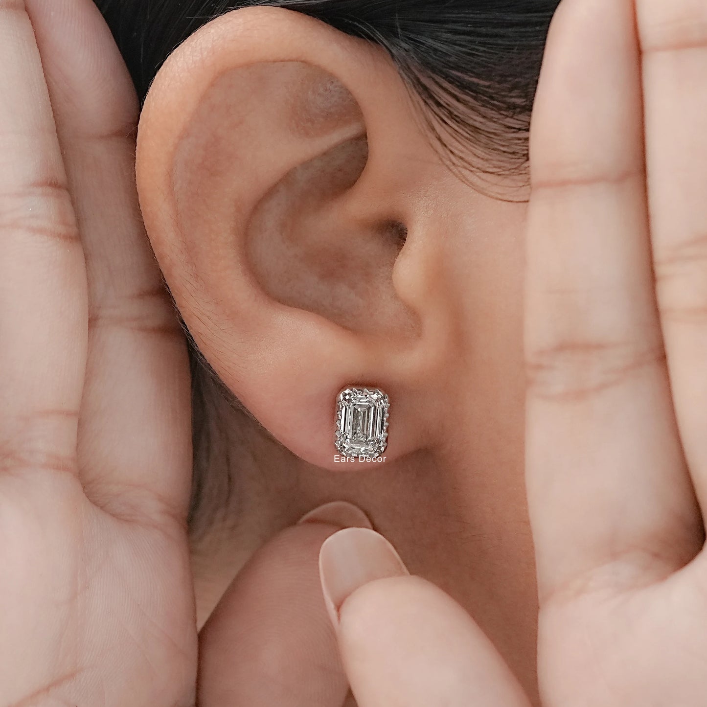 White gold emerald cut diamond studs worn on model's ear, both hands framing close-up