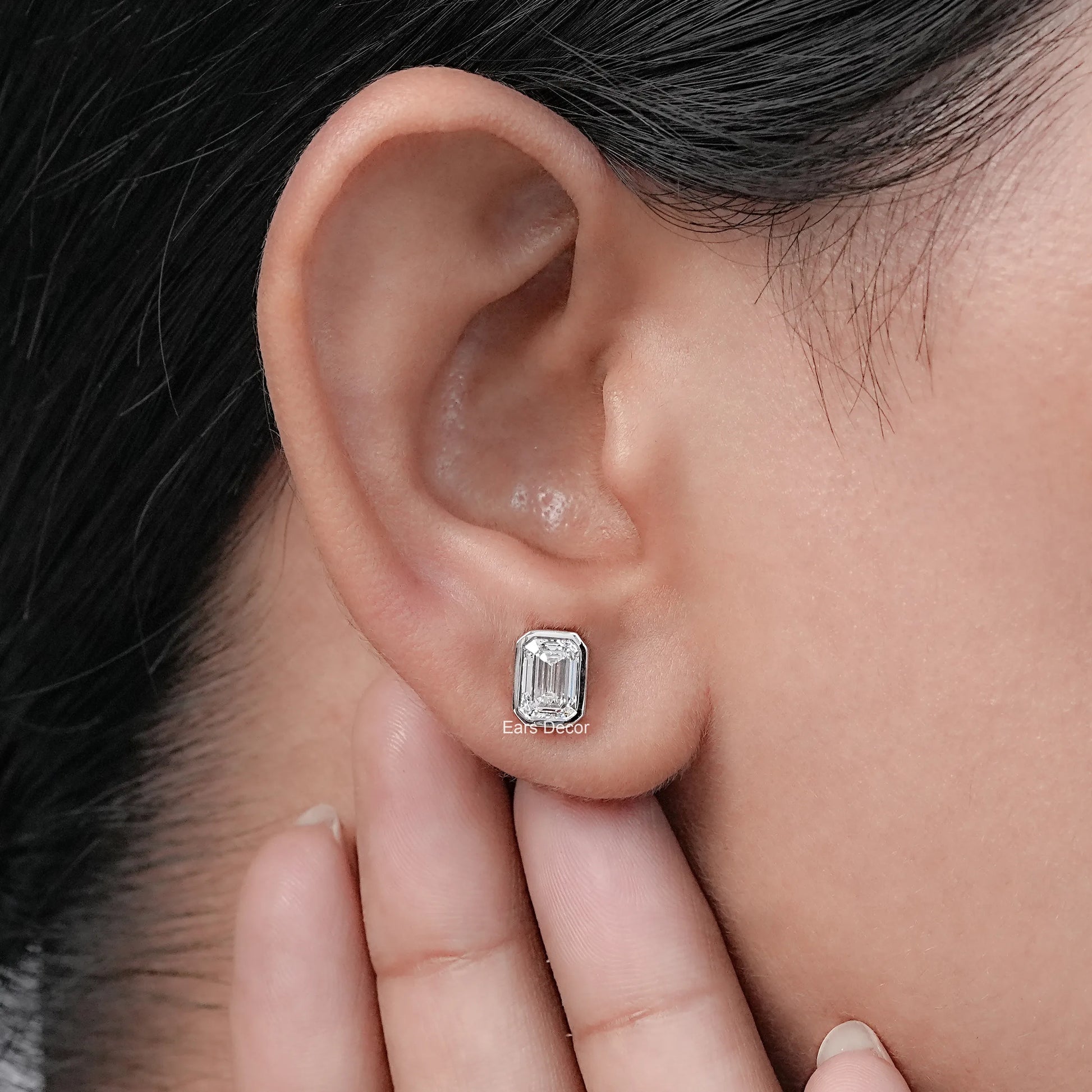 Emerald cut diamond stud earrings white gold bezel setting worn model ear hand touching dark hair warm skin close-up side profile