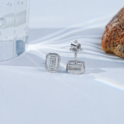 Emerald cut solitaire diamond stud earrings white gold bezel modern minimalist pair flat lay white linen side angle glass coral decor