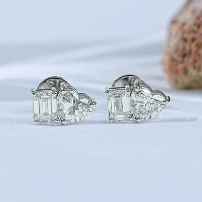 White gold toi et moi diamond stud earrings heart and emerald cut pair flat lay white satin fabric coral rock background close-up