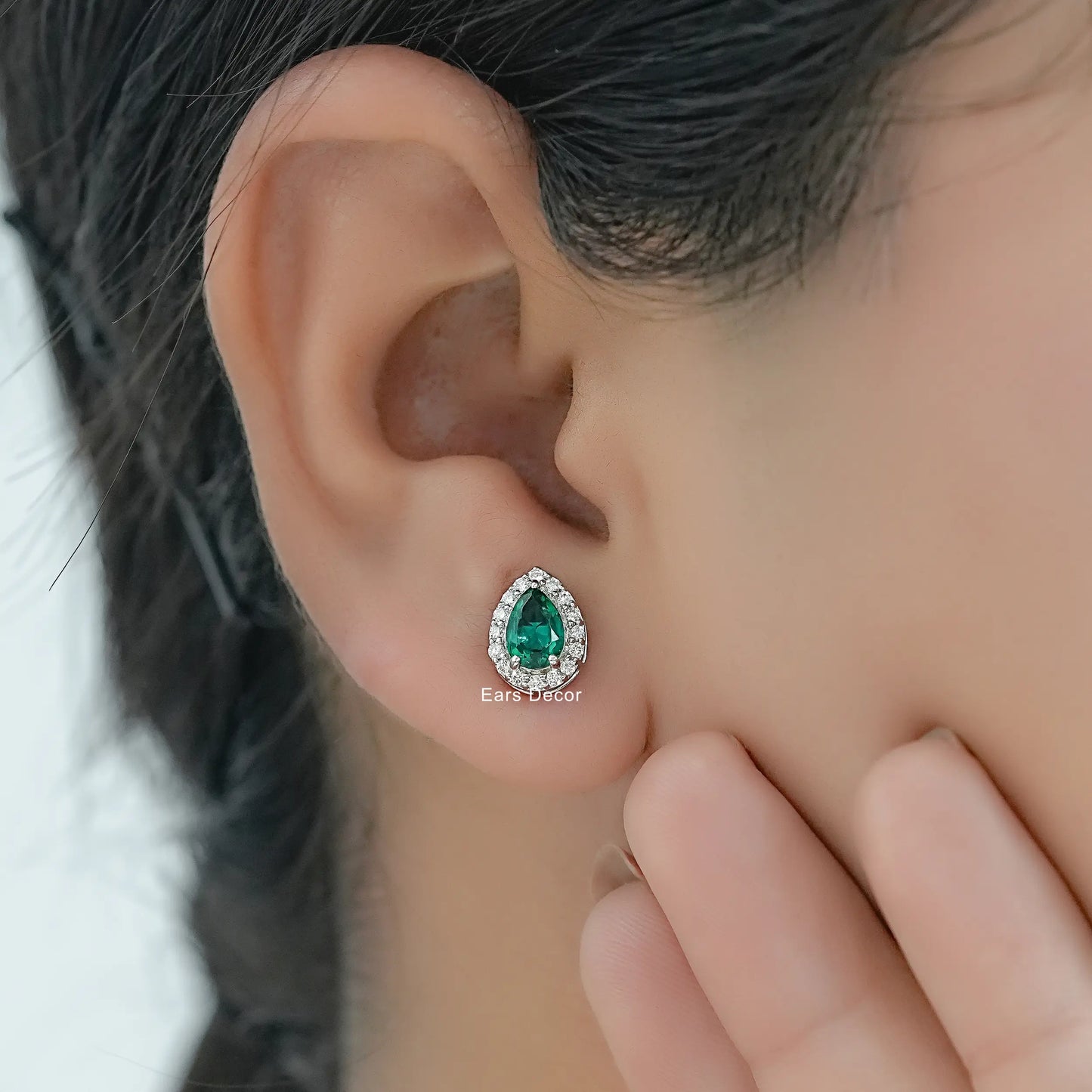 Lab created emerald halo stud earrings with brilliant cut diamond accents worn on model ear close-up dark hair hand touching face
