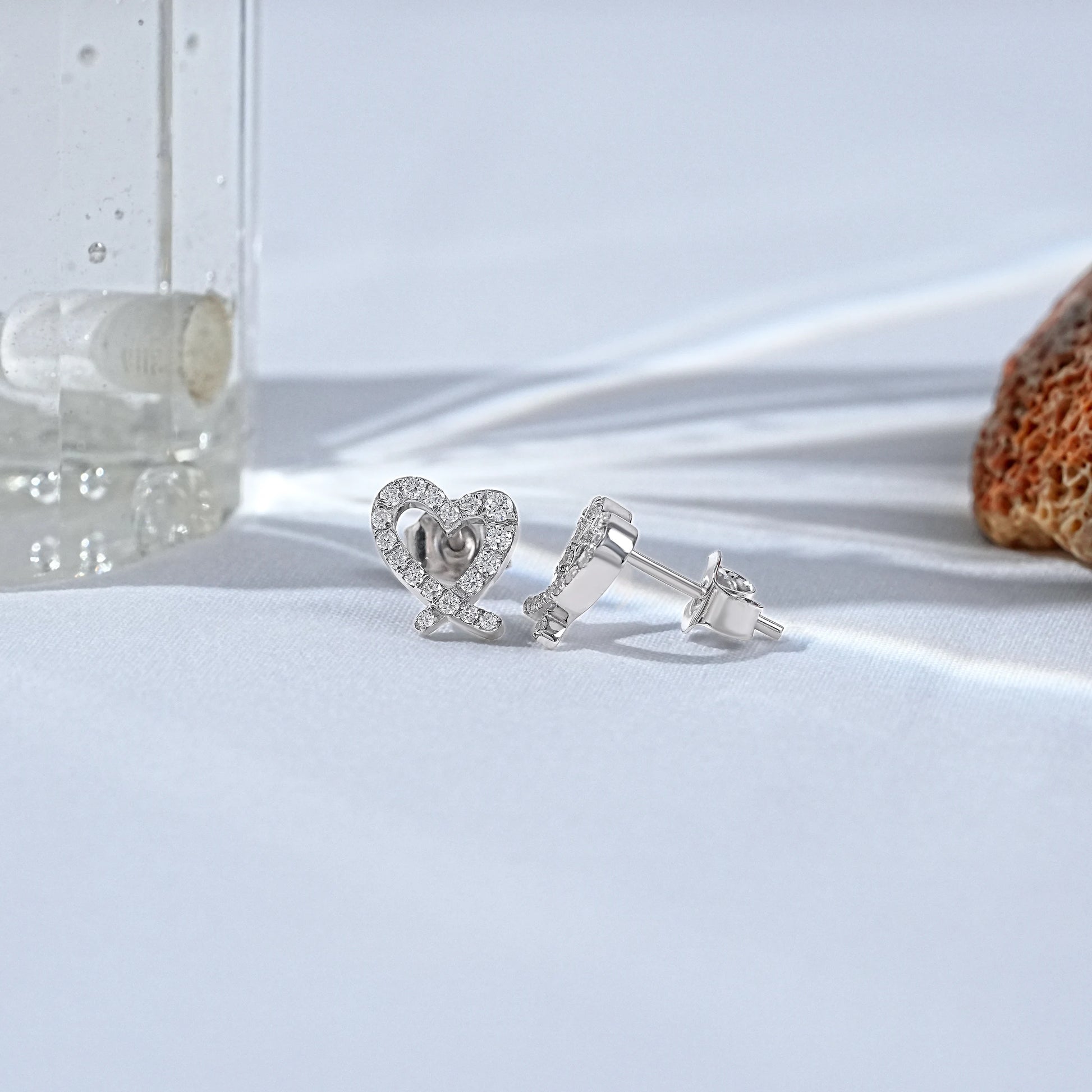 Minimalist heart shaped diamond earrings in white gold displayed on a clean, bright studio surface.
