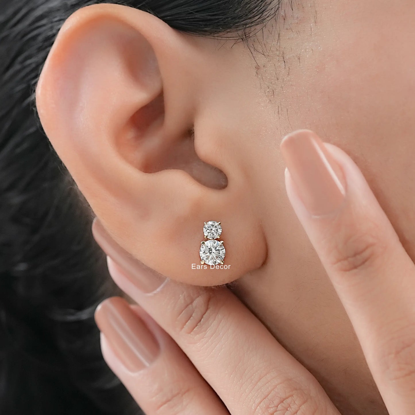 Dainty double diamond earrings yellow gold round brilliant stones worn on model ear close-up dark hair fair skin nude mauve nails fingers touching cheek
