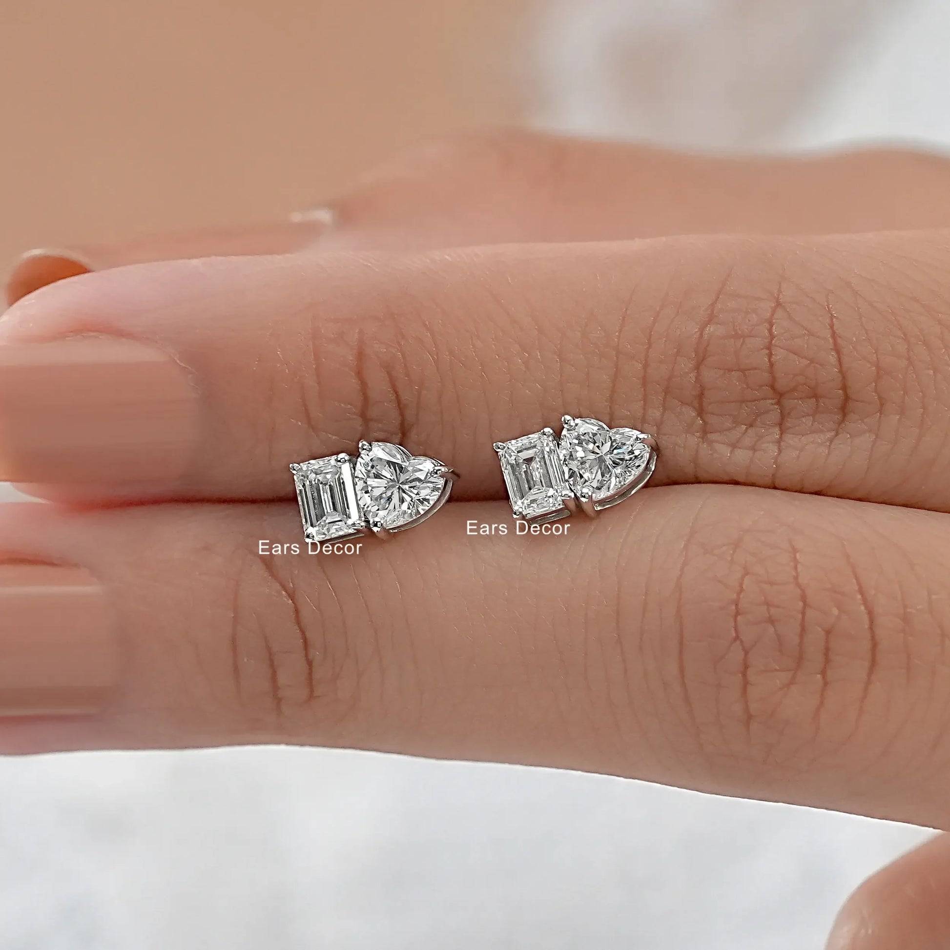 Romantic heart and emerald diamond earrings gift girlfriend wife white gold pair held on warm skin model fingers close-up nude pink nails peach background
