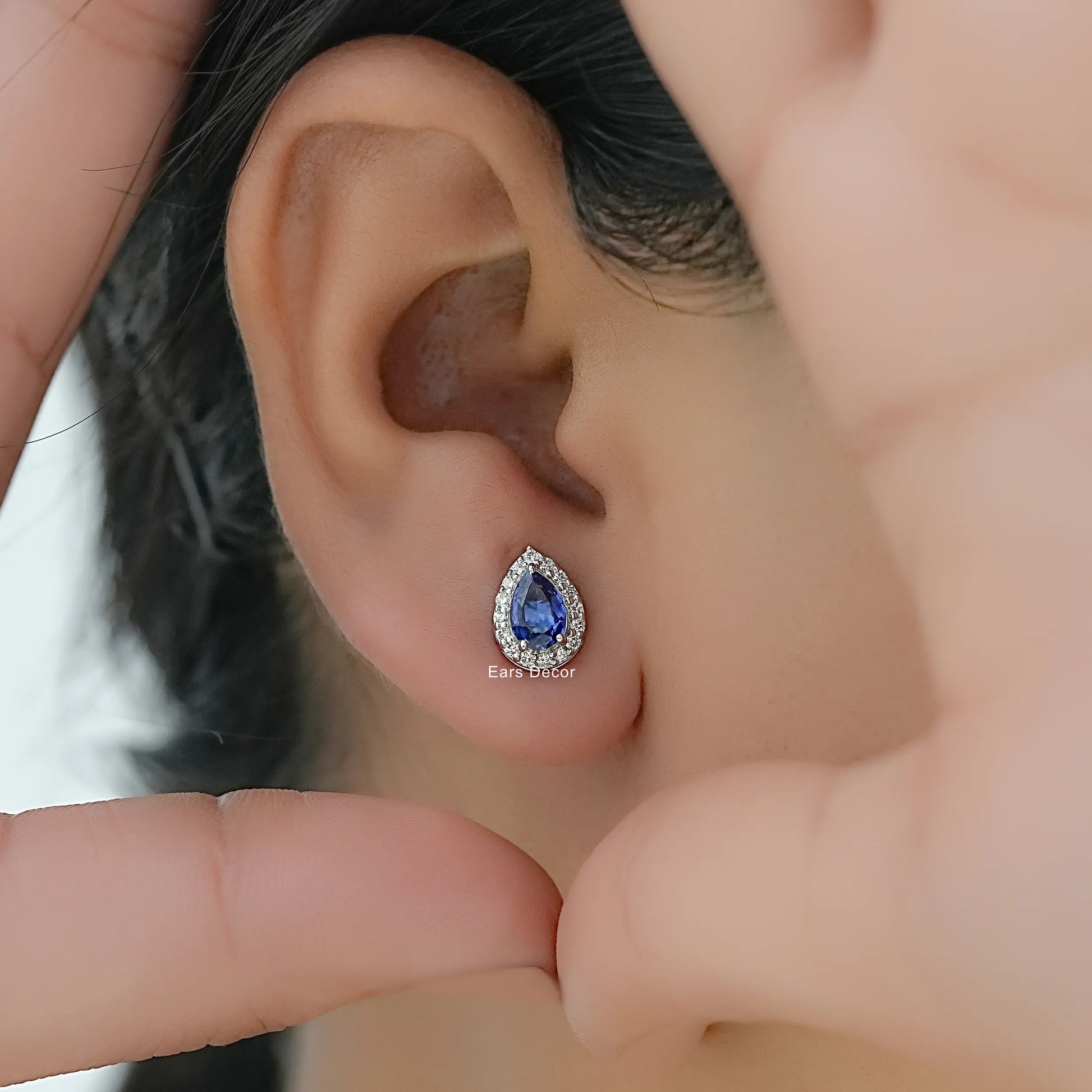 Pear shaped sapphire and diamond cluster stud earrings worn on model ear close-up dark hair fingers framing face warm skin
