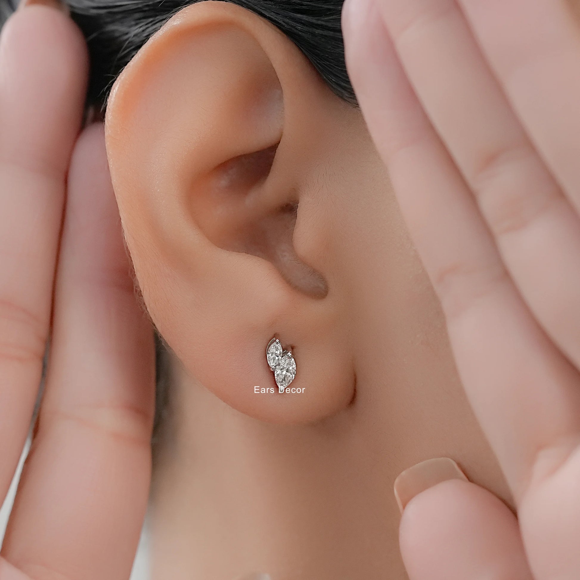 Small dainty toi et moi diamond studs cartilage or lobe worn on model ear close-up dark hair both hands framing face warm skin