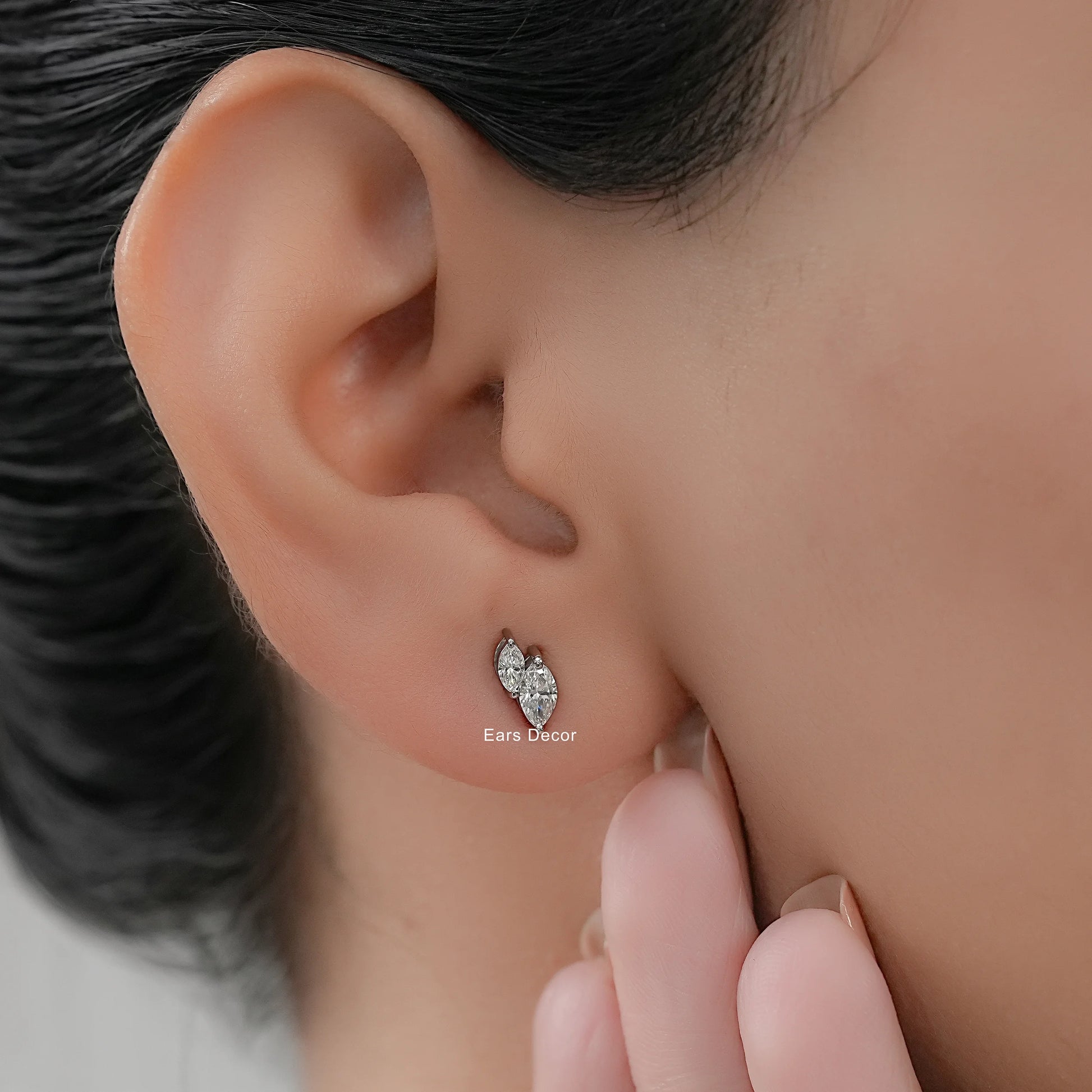 Toi et moi diamond studs marquise cut stones worn on model ear close-up dark hair nude nails hand touching cheek warm skin
