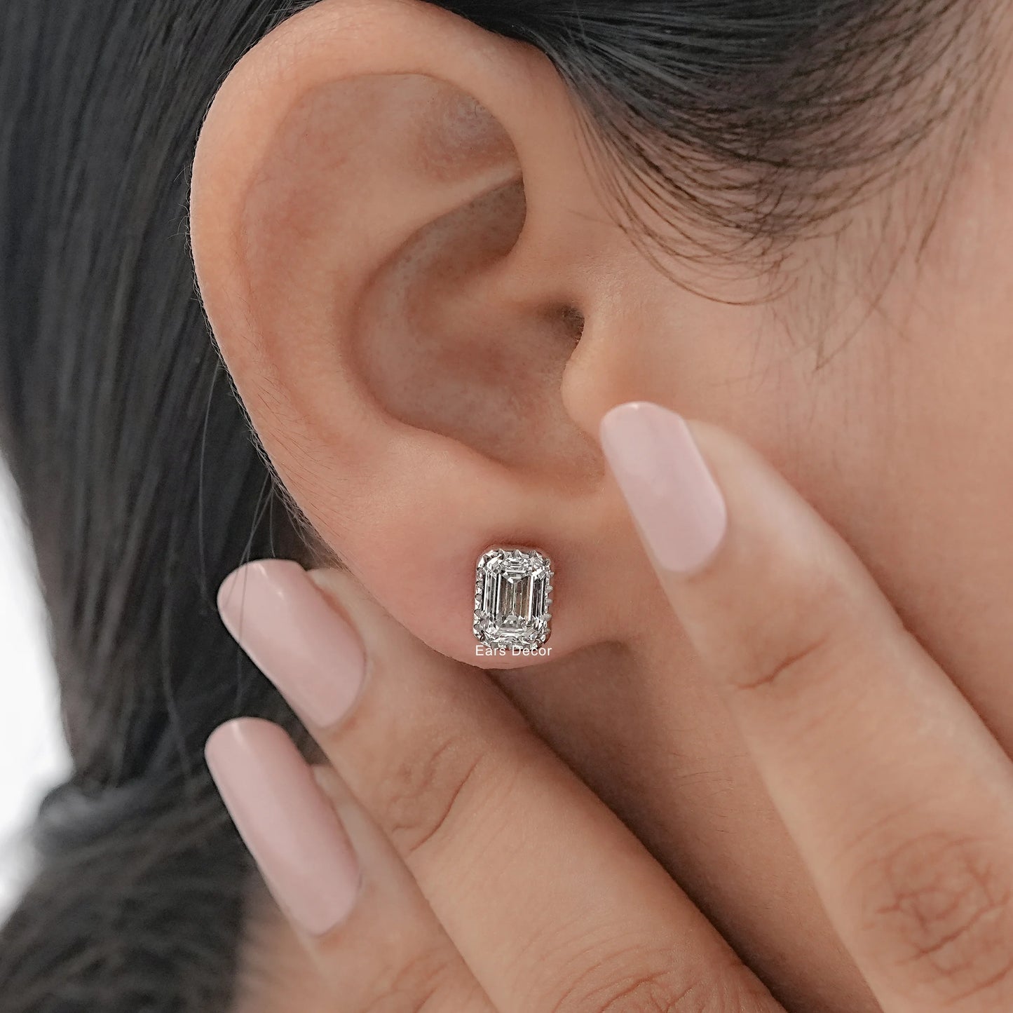 Emerald cut solitaire diamond stud earring worn on model's ear with nude pink manicure