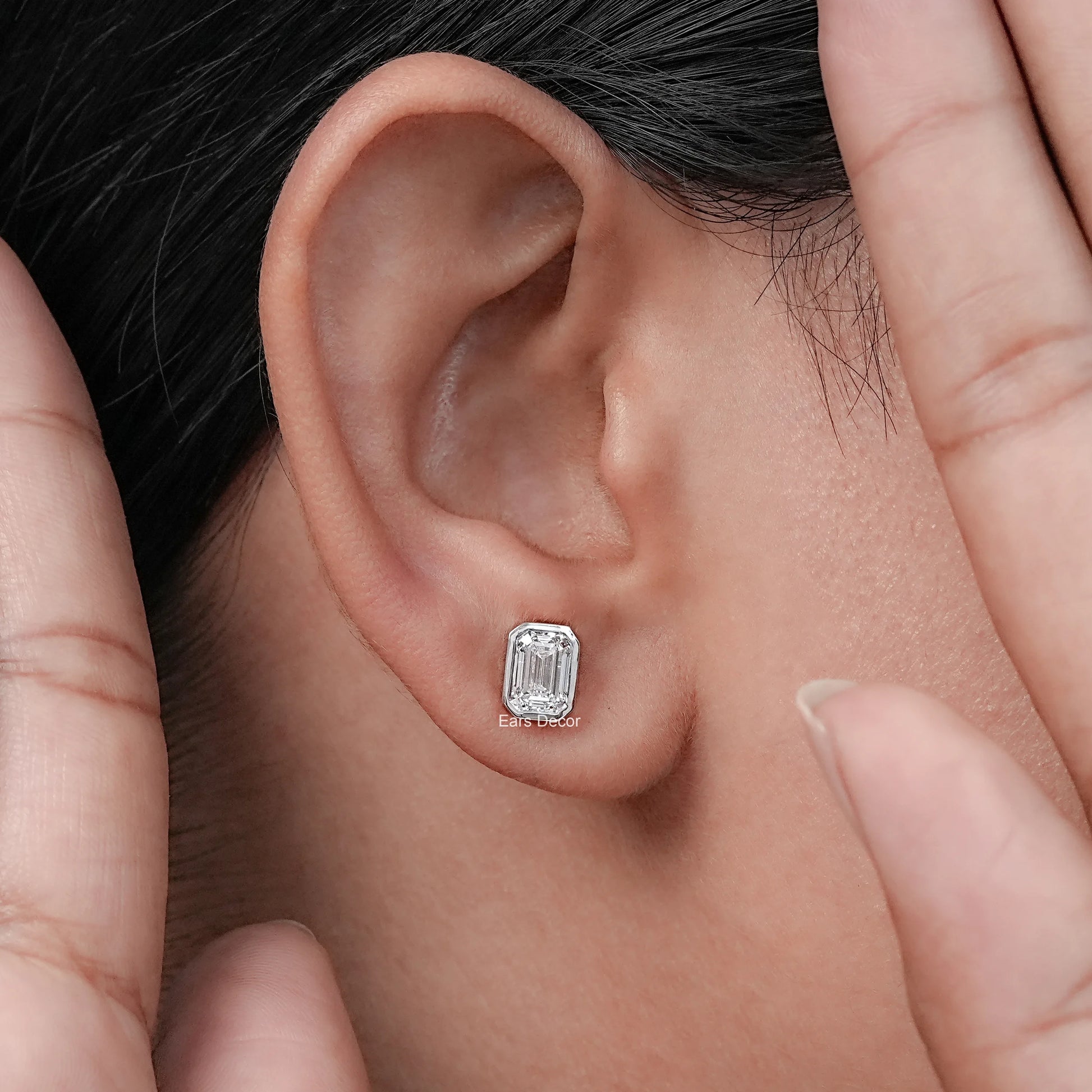 Emerald cut diamond stud earrings geometric design white gold bezel worn model ear both hands framing dark hair warm skin close-up