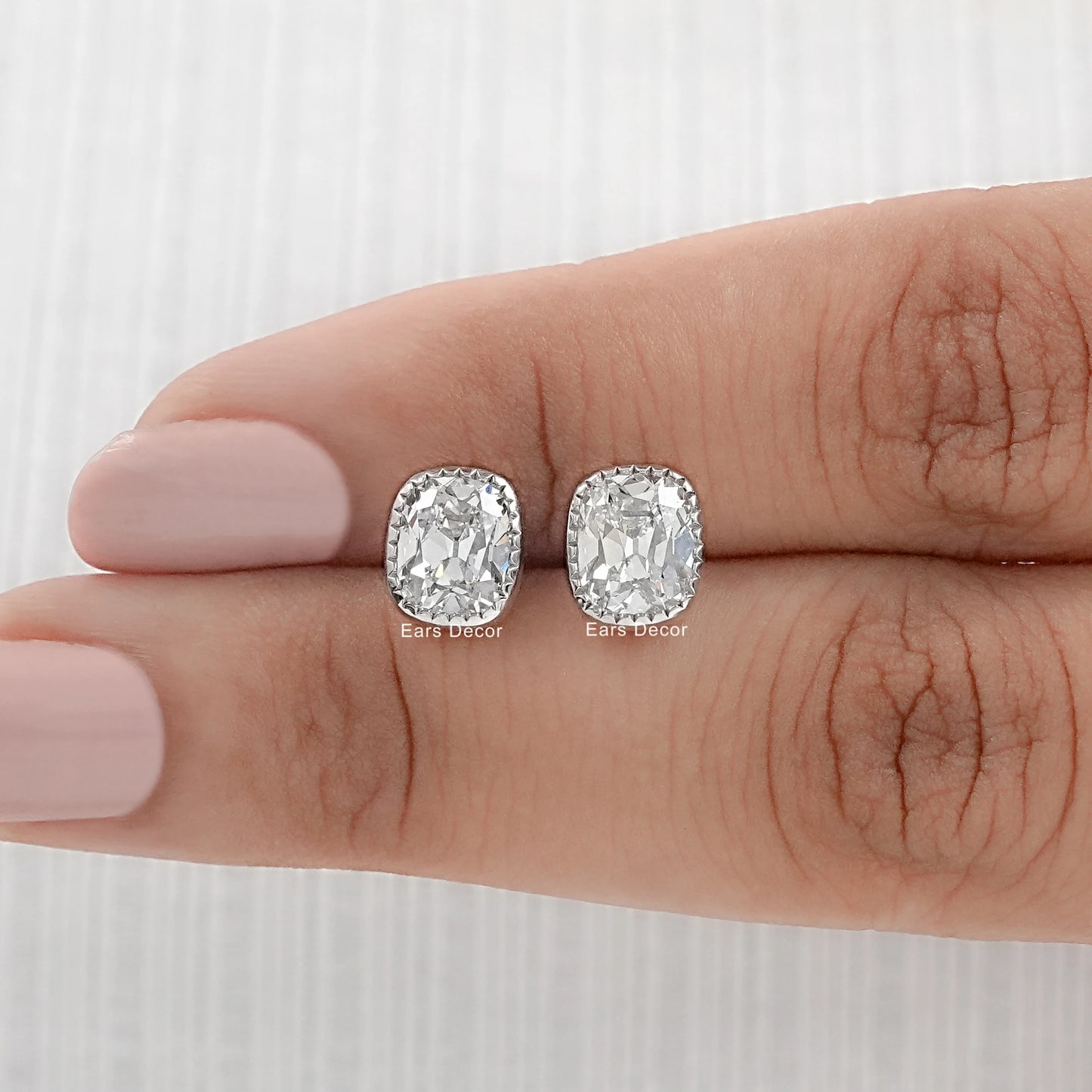 Minimalist cushion cut diamond stud earrings white gold milgrain bezel pair displayed on fingers dark skin nude manicure close-up