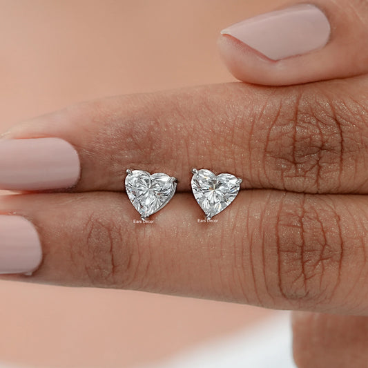 A pair of heart-shaped diamond solitaire stud earrings placed on a person's finger, showcasing the earrings' design and the clarity of the diamonds.
