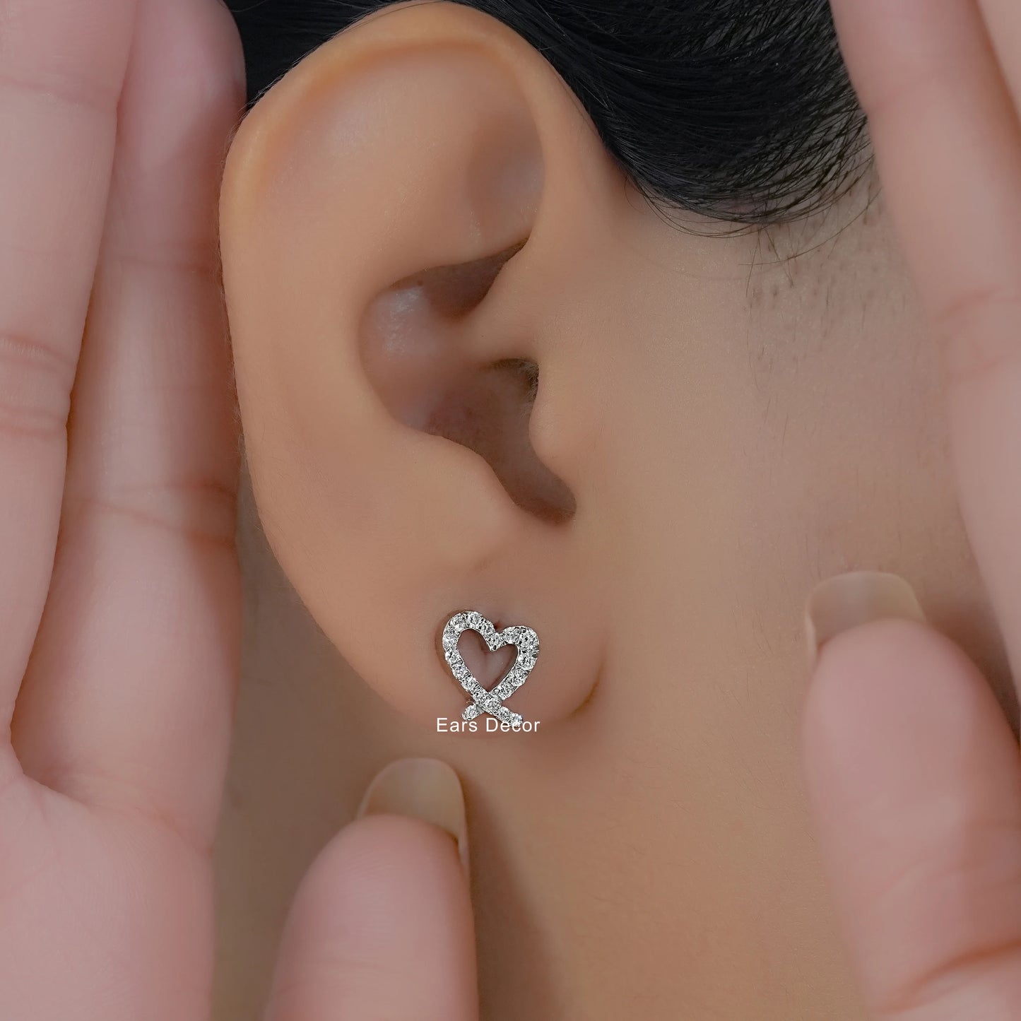 White gold pavé heart diamond earrings worn on a model ear framed by hands with neutral nails.