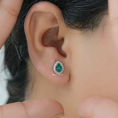 Pear cut emerald teardrop stud earrings solid white gold diamond halo worn on model ear close-up dark hair fingers visible