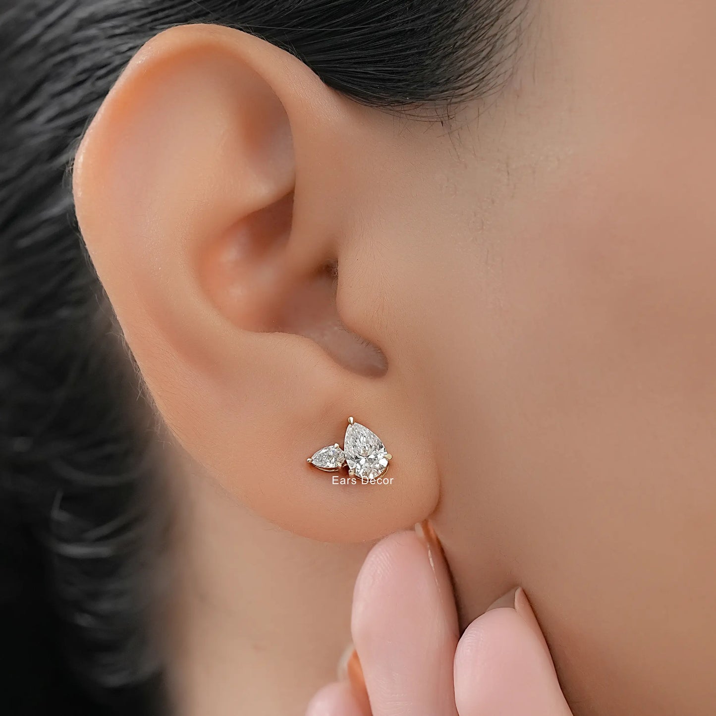Minimalist pear shaped diamond stud earrings with side stones yellow gold worn on model ear close-up dark hair nude nails fingers touching jaw