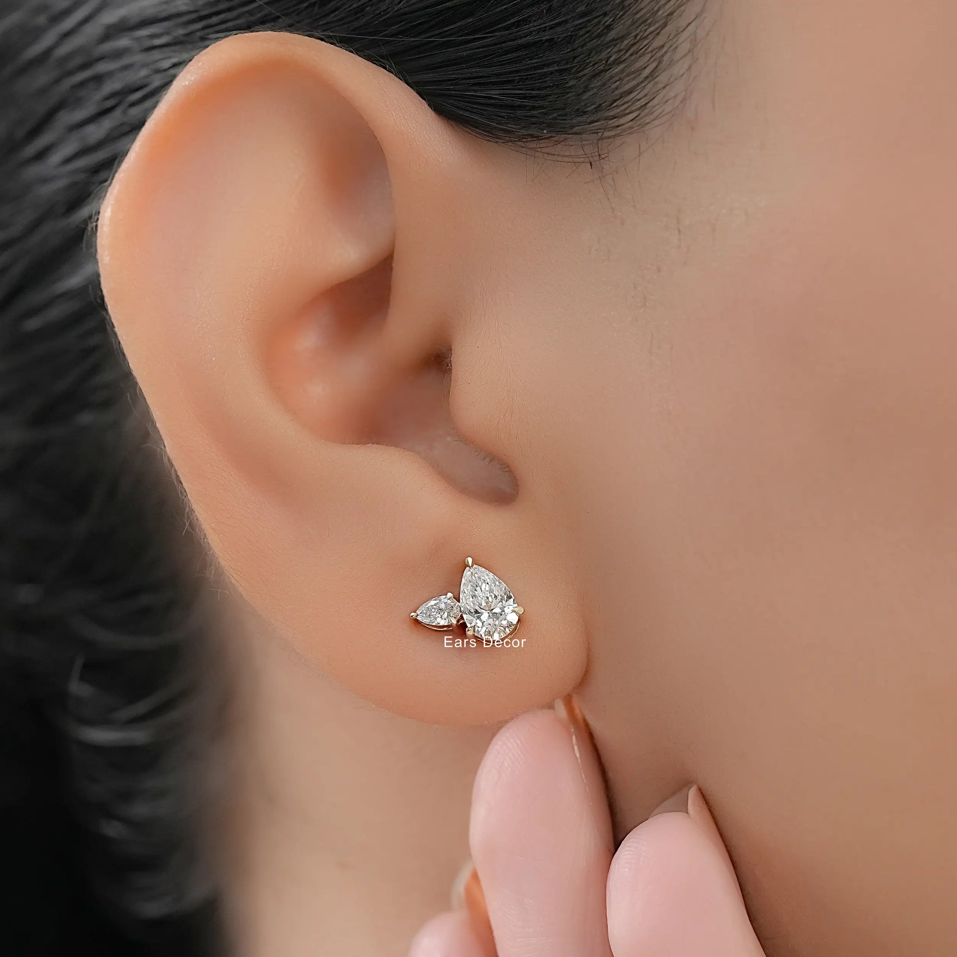 Minimalist pear shaped diamond stud earrings with side stones yellow gold worn on model ear close-up dark hair nude nails fingers touching jaw