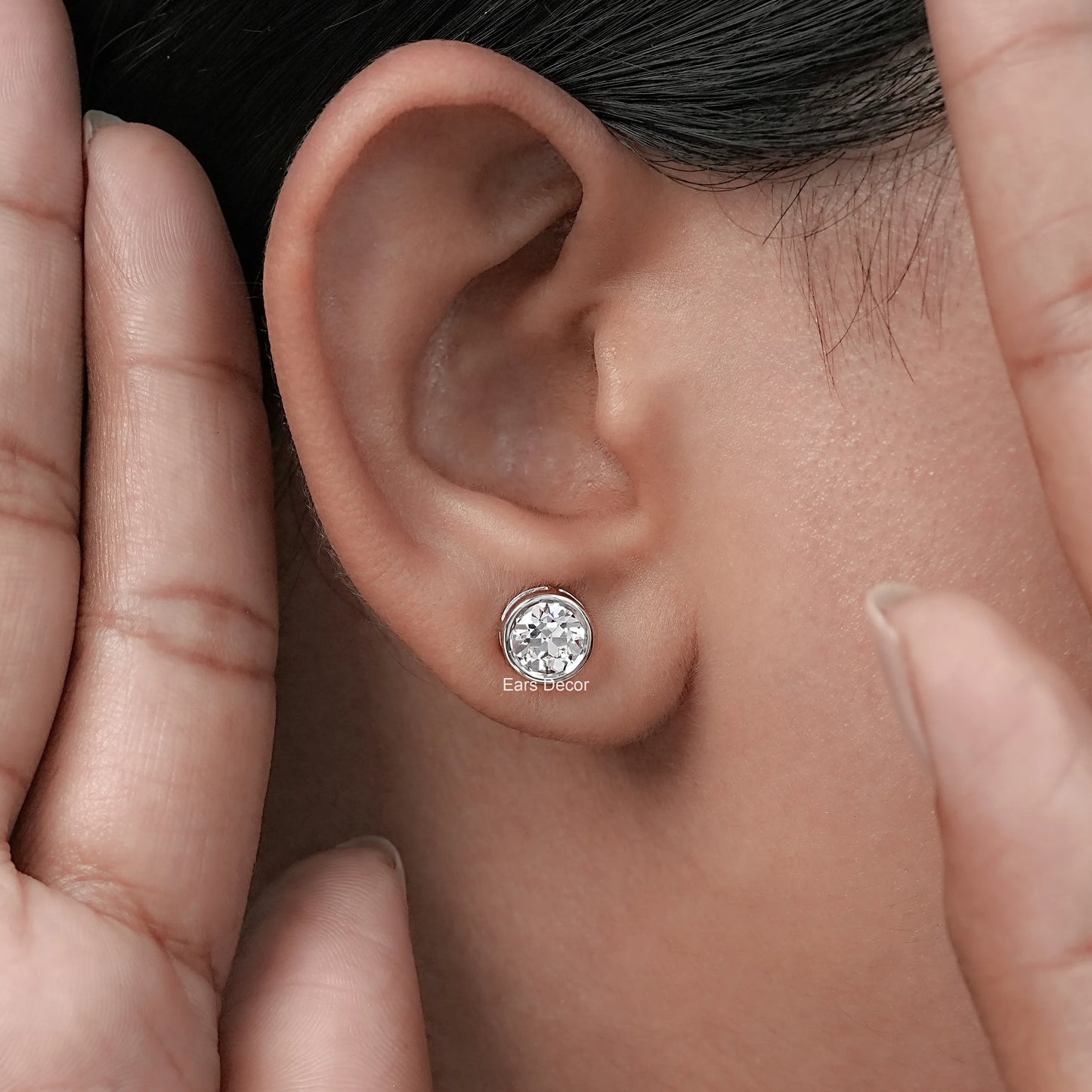 Old European round cut lab diamond bezel set stud earrings worn in a woman's ear with a hand gently touching her ear.