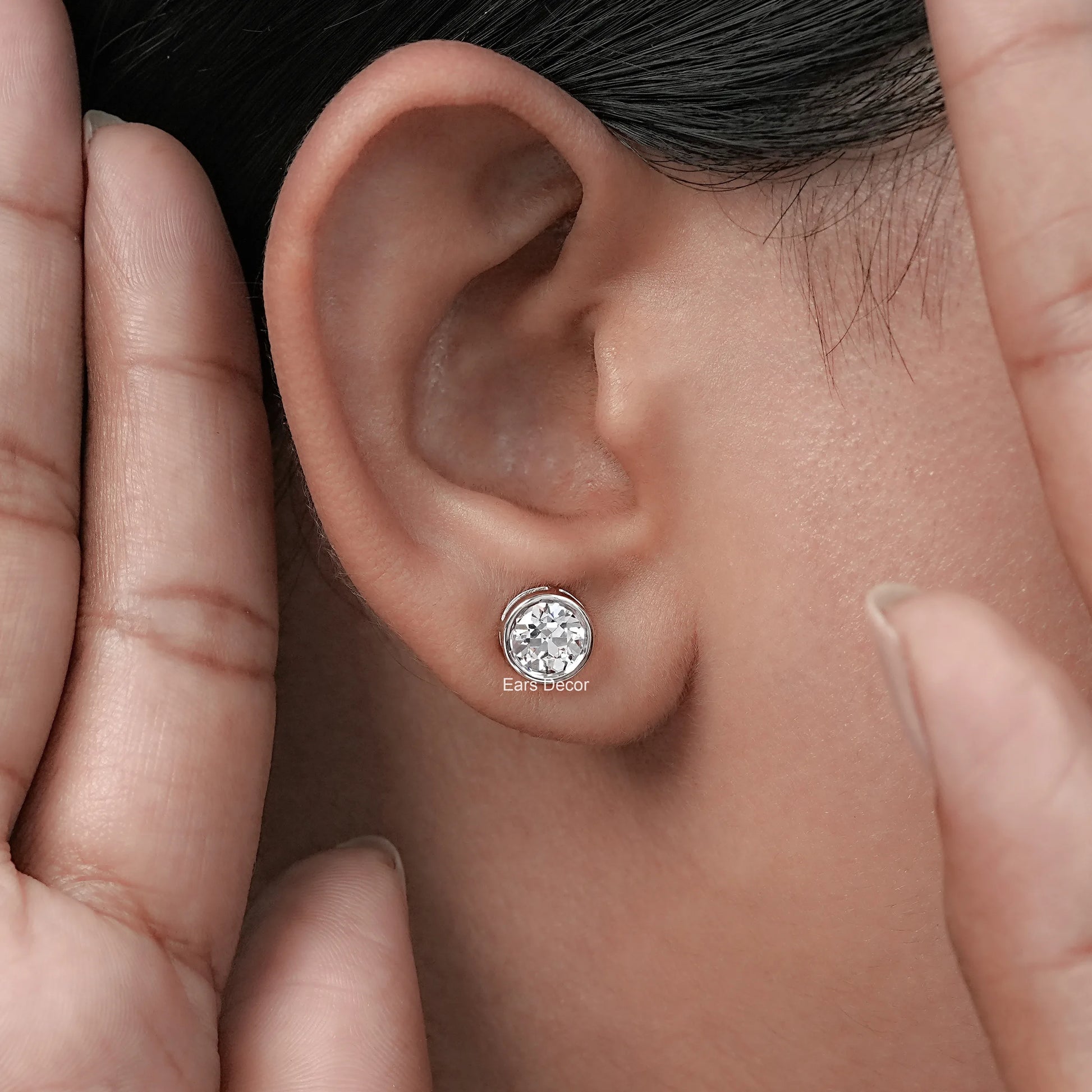 Old European round cut lab diamond bezel set stud earrings worn in a woman's ear with a hand gently touching her ear.