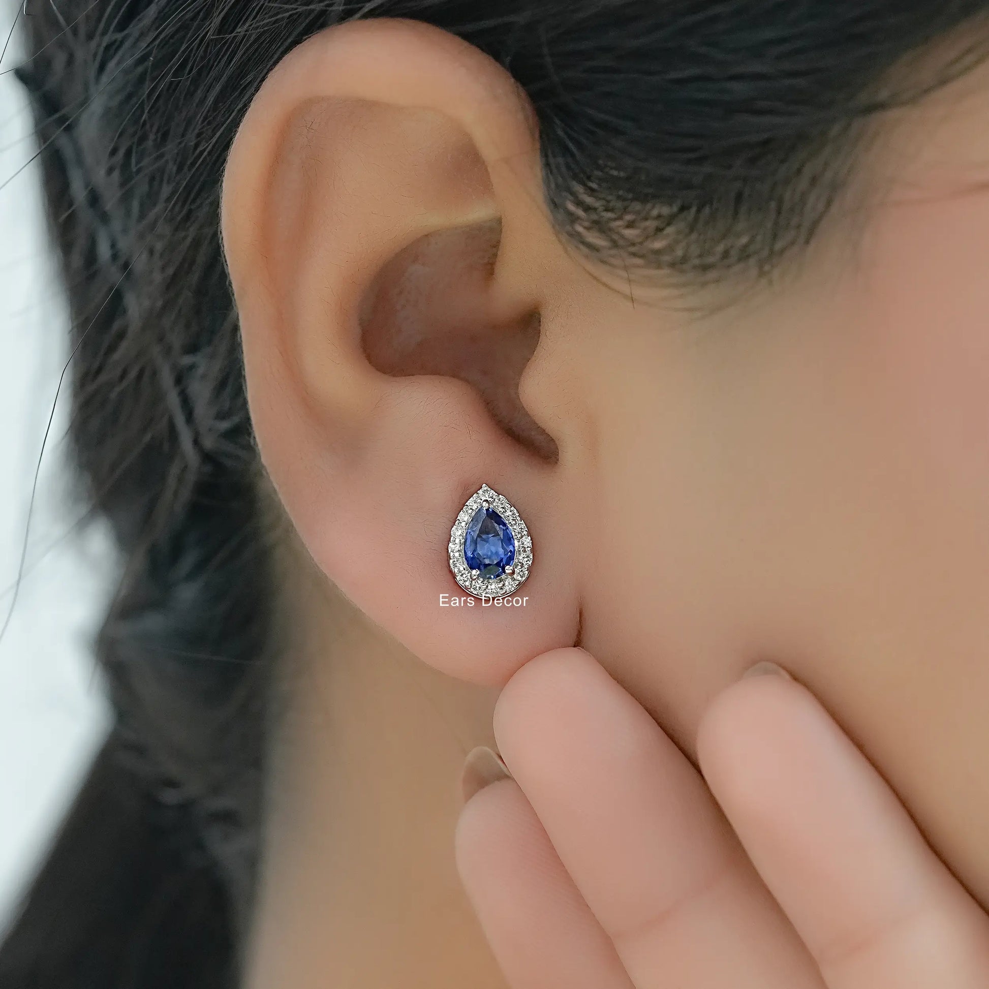 Blue sapphire teardrop halo earrings with round diamond accents worn on model ear close-up dark braided hair hand touching face