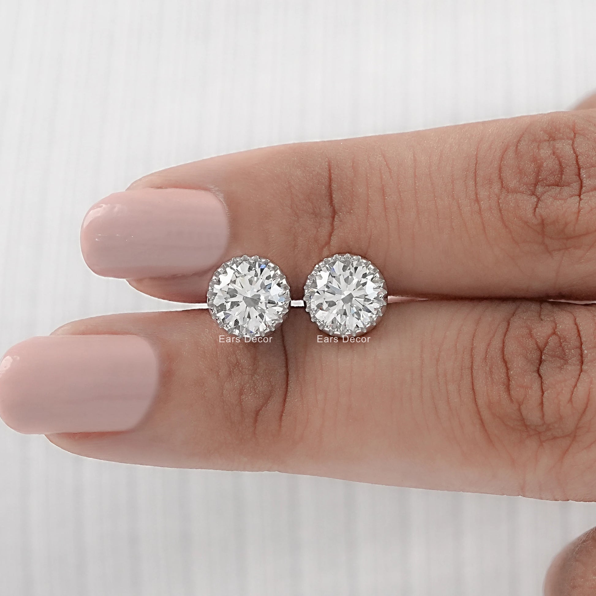 Round cut moissanite stud earrings with martini setting white gold pair displayed on model fingers nude nails close-up