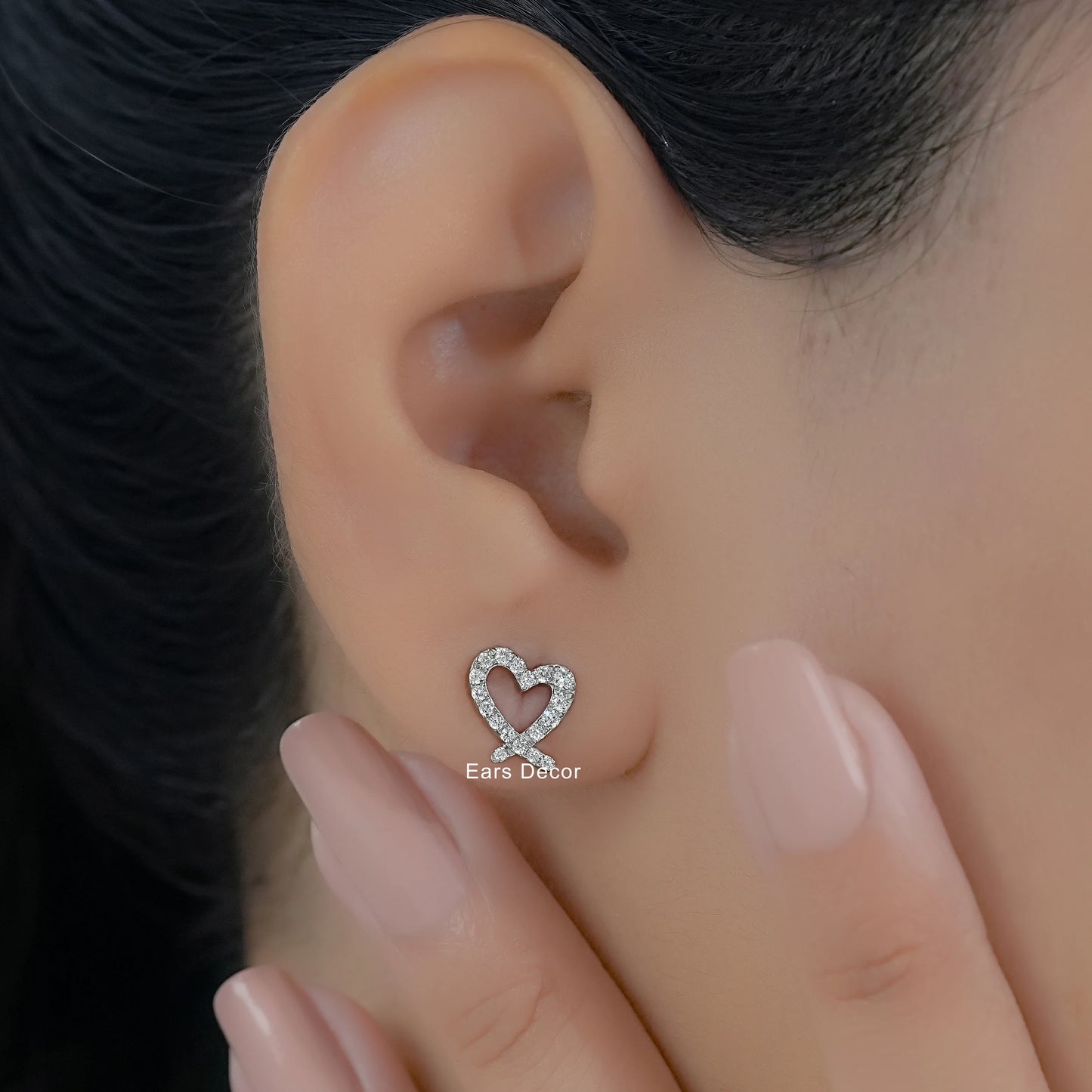 Minimalist heart shaped diamond earrings worn on a model with a soft-focus background and nude manicure.