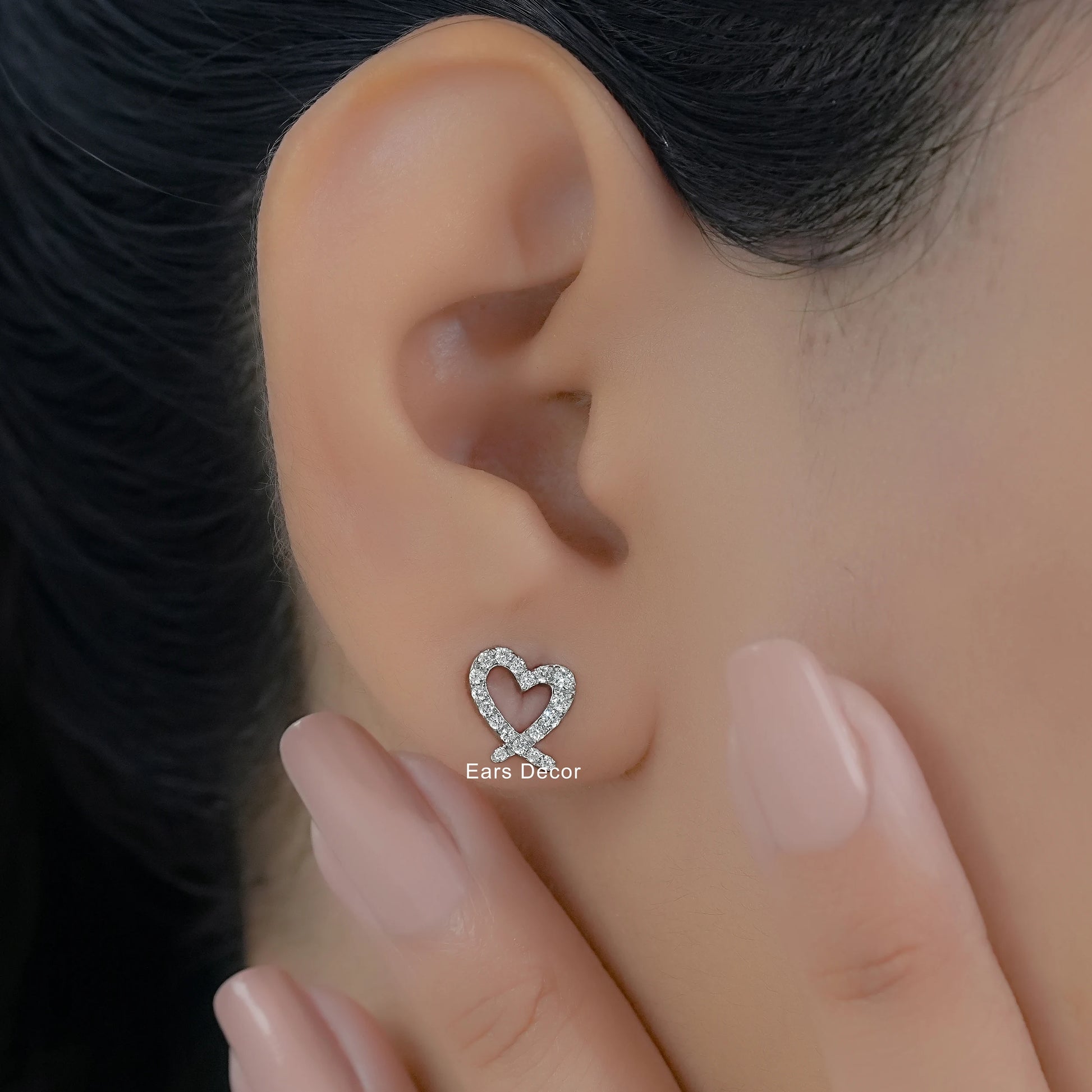 Minimalist heart shaped diamond earrings worn on a model with a soft-focus background and nude manicure.