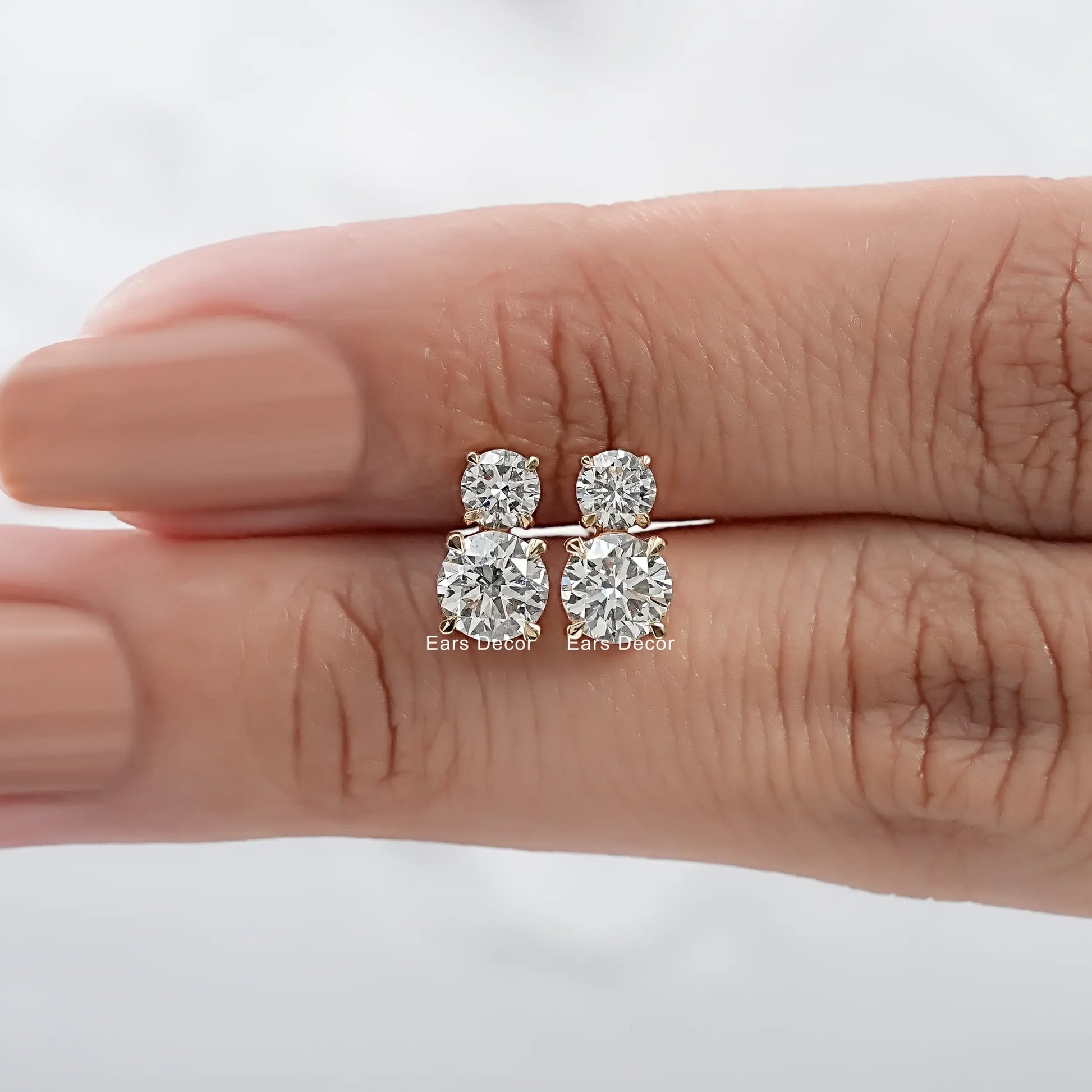 Yellow gold double diamond stud earrings two round brilliant stones pair held on warm skin model fingers close-up nude mauve nails white background