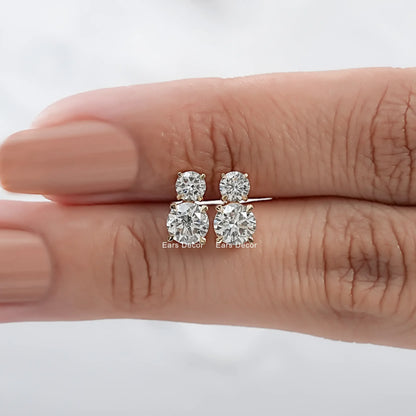 Yellow gold double diamond stud earrings two round brilliant stones pair held on warm skin model fingers close-up nude mauve nails white background