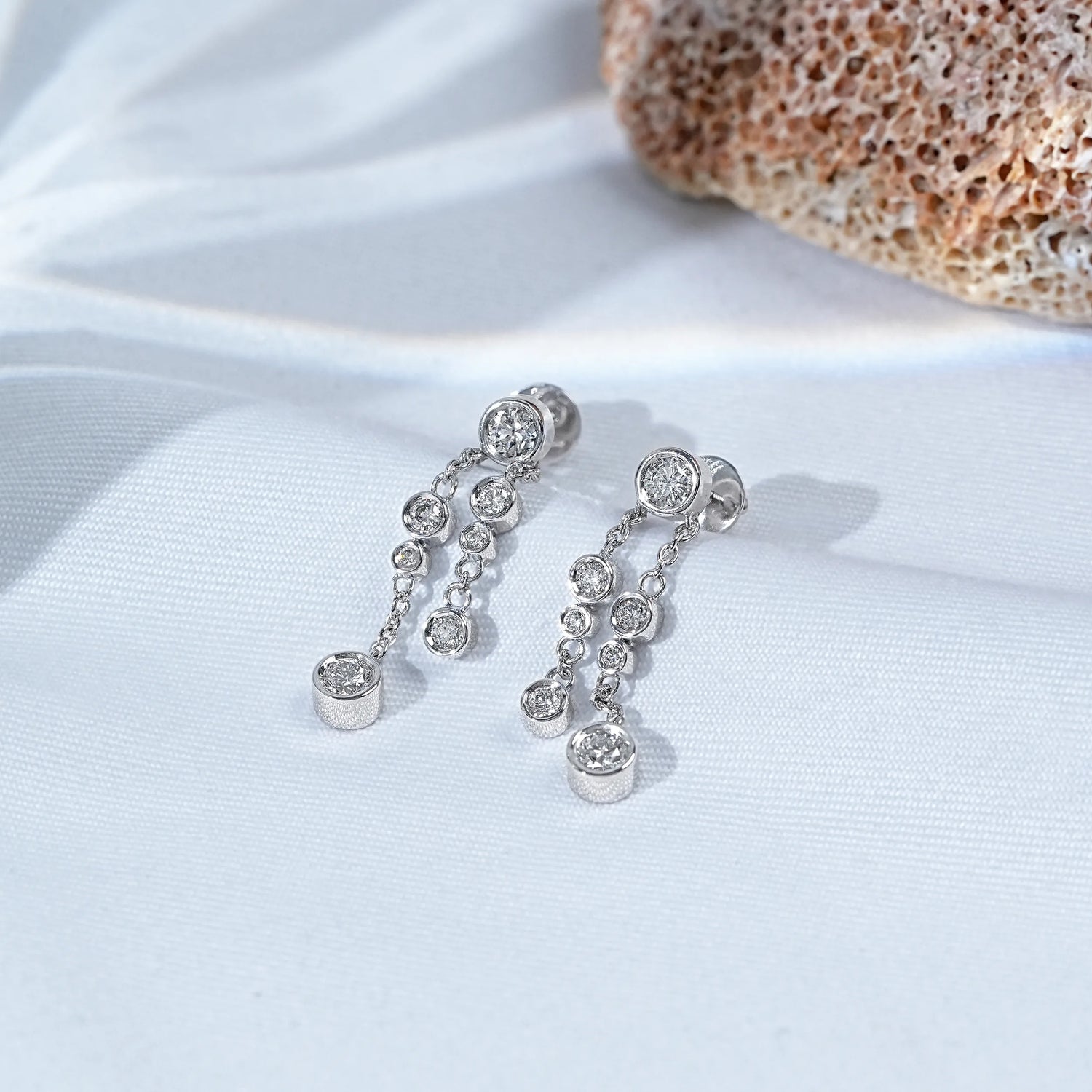 Round bezel set chain dangle drop earrings laid on white satin beside coral rock prop full pair view