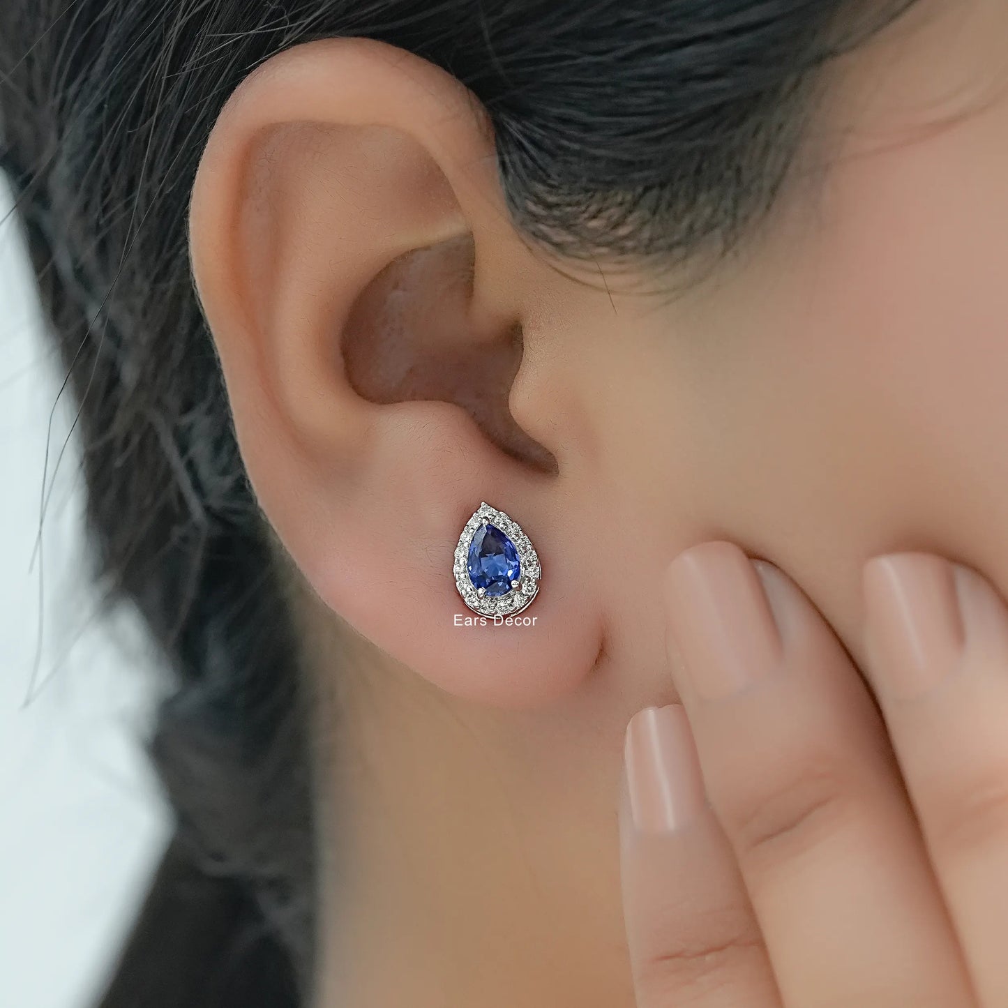Minimalist pear shaped sapphire halo cluster stud earrings worn on model ear close-up dark hair nude nails hand touching cheek