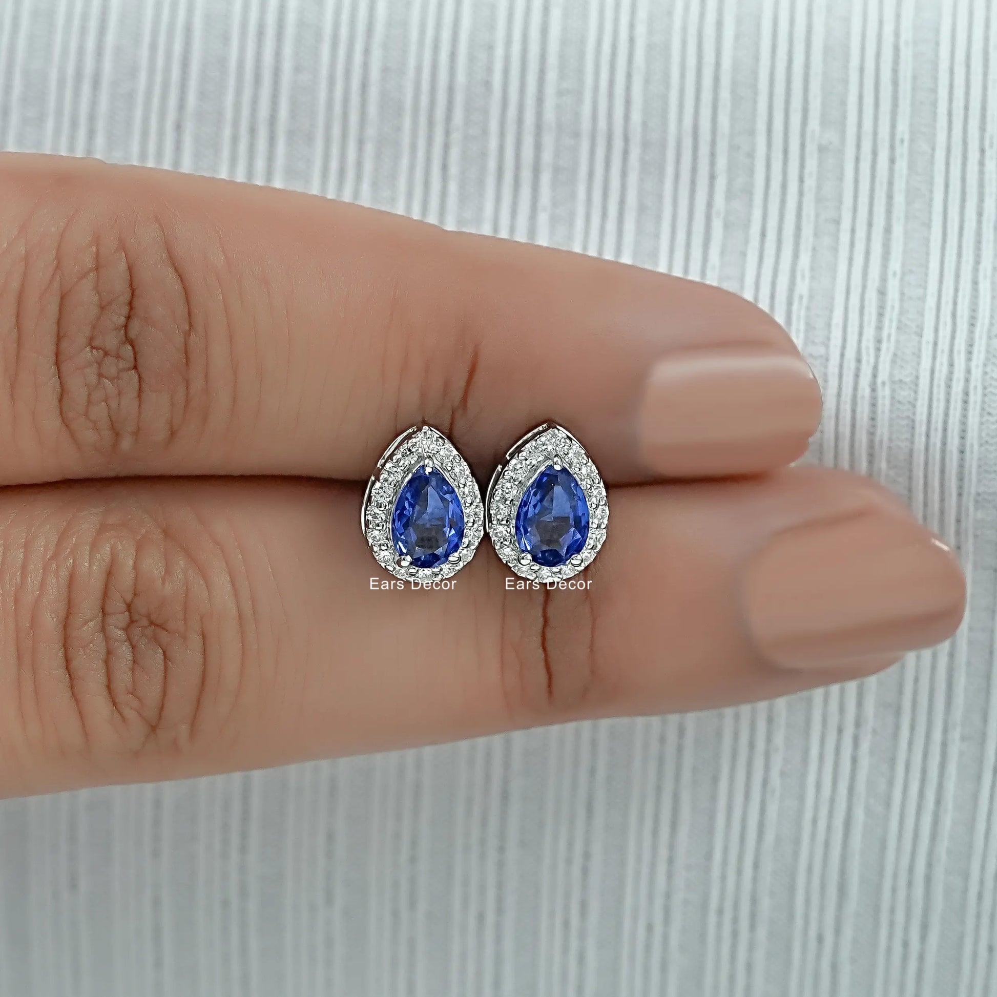 Teardrop blue sapphire stud earrings white gold diamond halo held between two fingers size reference nude nails close-up warm skin
