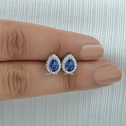 Teardrop blue sapphire stud earrings white gold diamond halo held between two fingers size reference nude nails close-up warm skin
