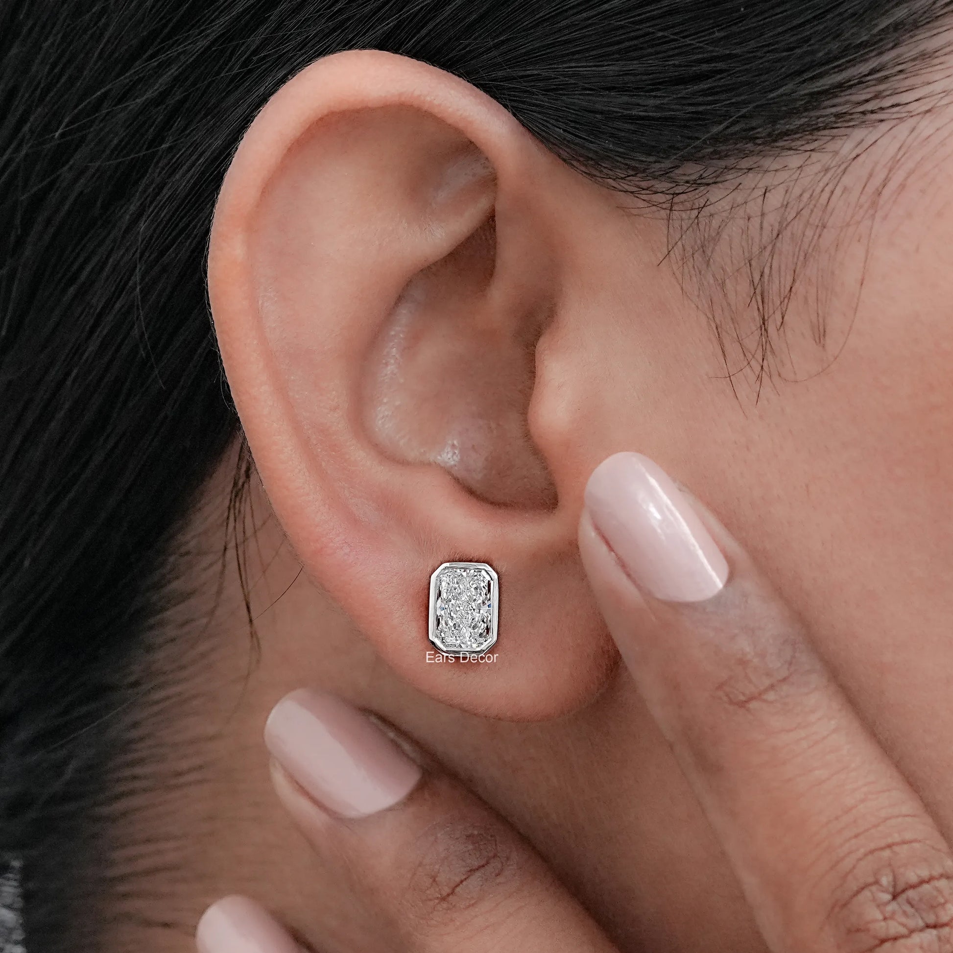 Octagon shaped bezel moissanite solitaire earring worn on model ear, nude nails fine jewelry