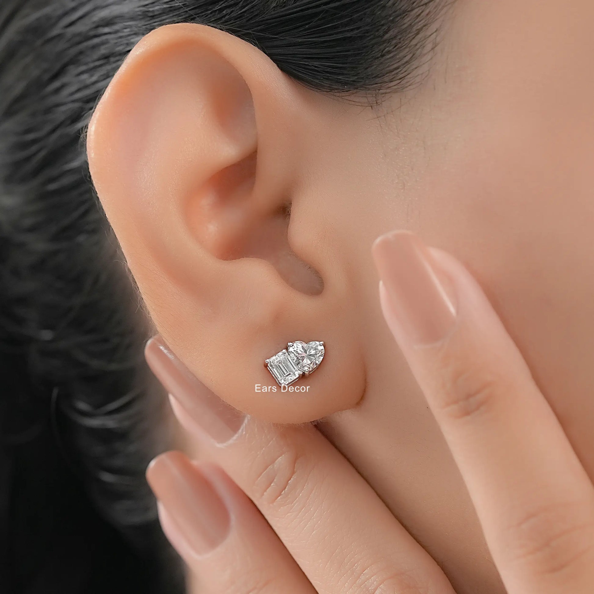 Dainty heart shaped and emerald cut diamond studs everyday wear white gold worn on model ear close-up dark hair warm skin nude mauve nails fingers touching