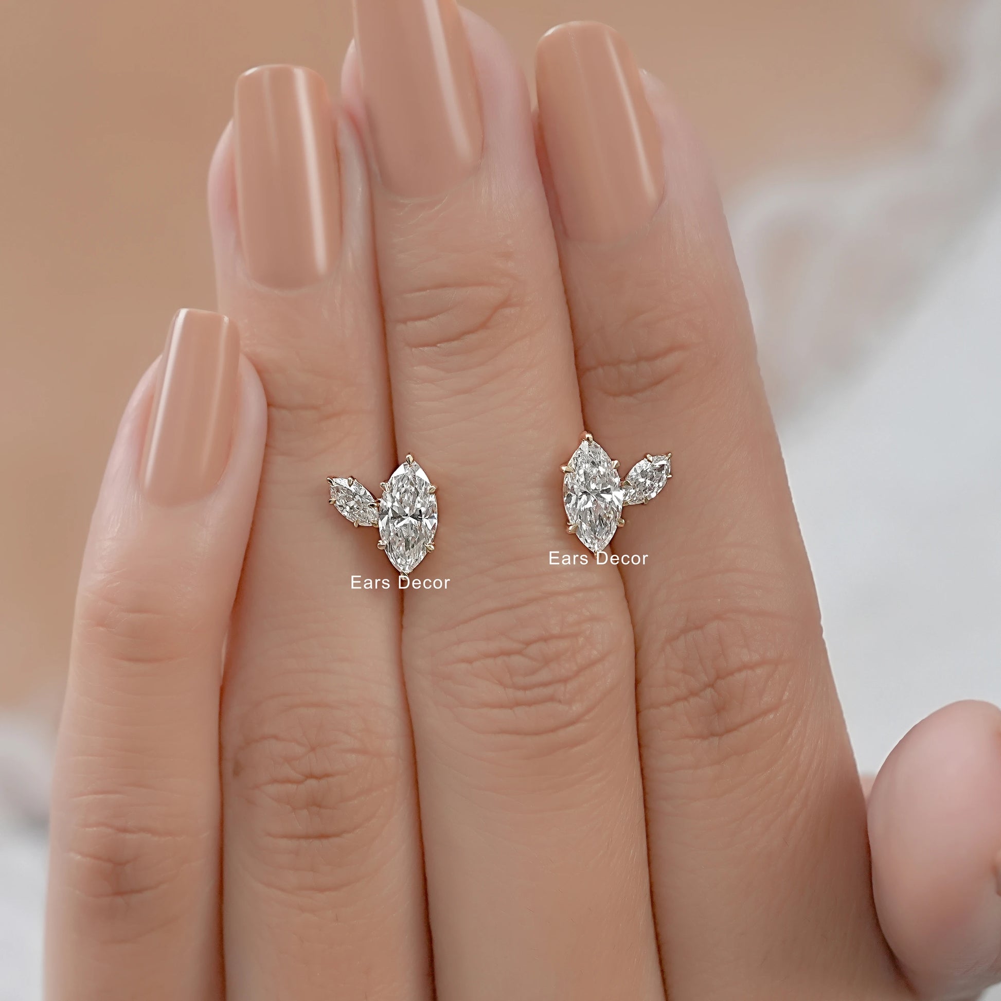 Marquise and pear cut diamond earrings with prong setting held between the fingers of a hand with nude nails.