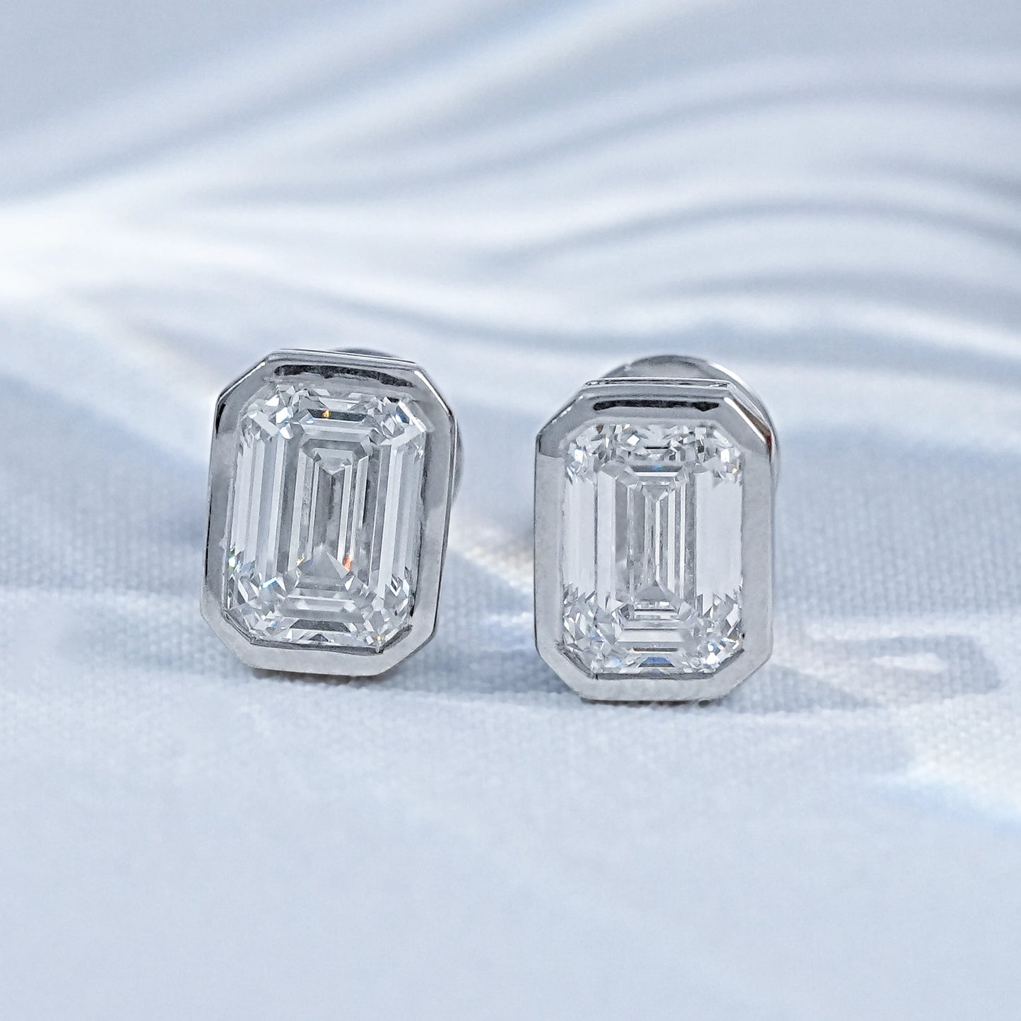 Emerald cut diamond stud earrings white gold bezel setting pair flat lay white linen soft light palm leaf shadow background