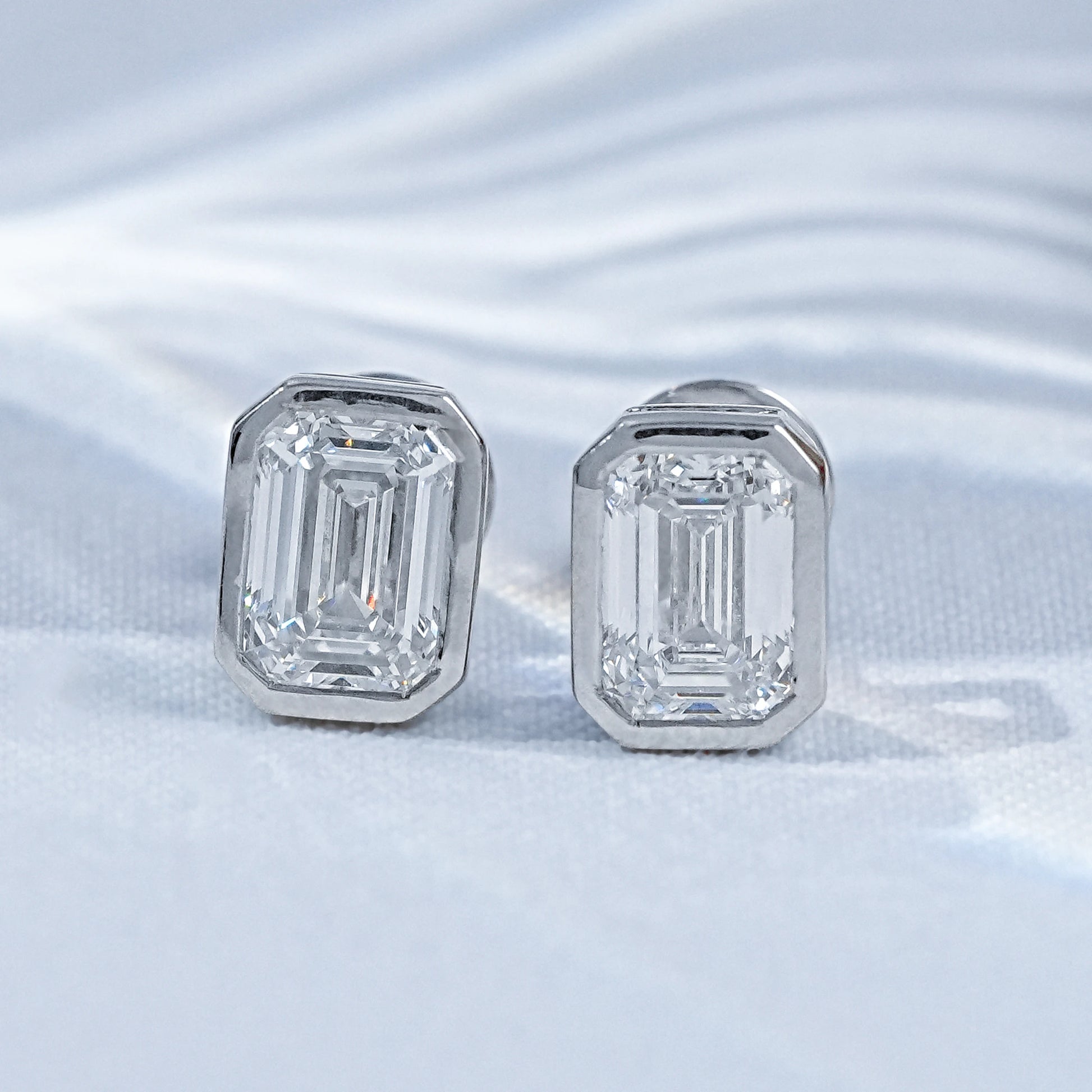 Emerald cut diamond stud earrings white gold bezel setting pair flat lay white linen soft light palm leaf shadow background