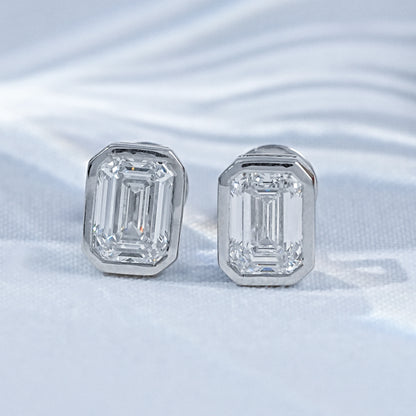 Emerald cut diamond stud earrings white gold bezel setting pair flat lay white linen soft light palm leaf shadow background